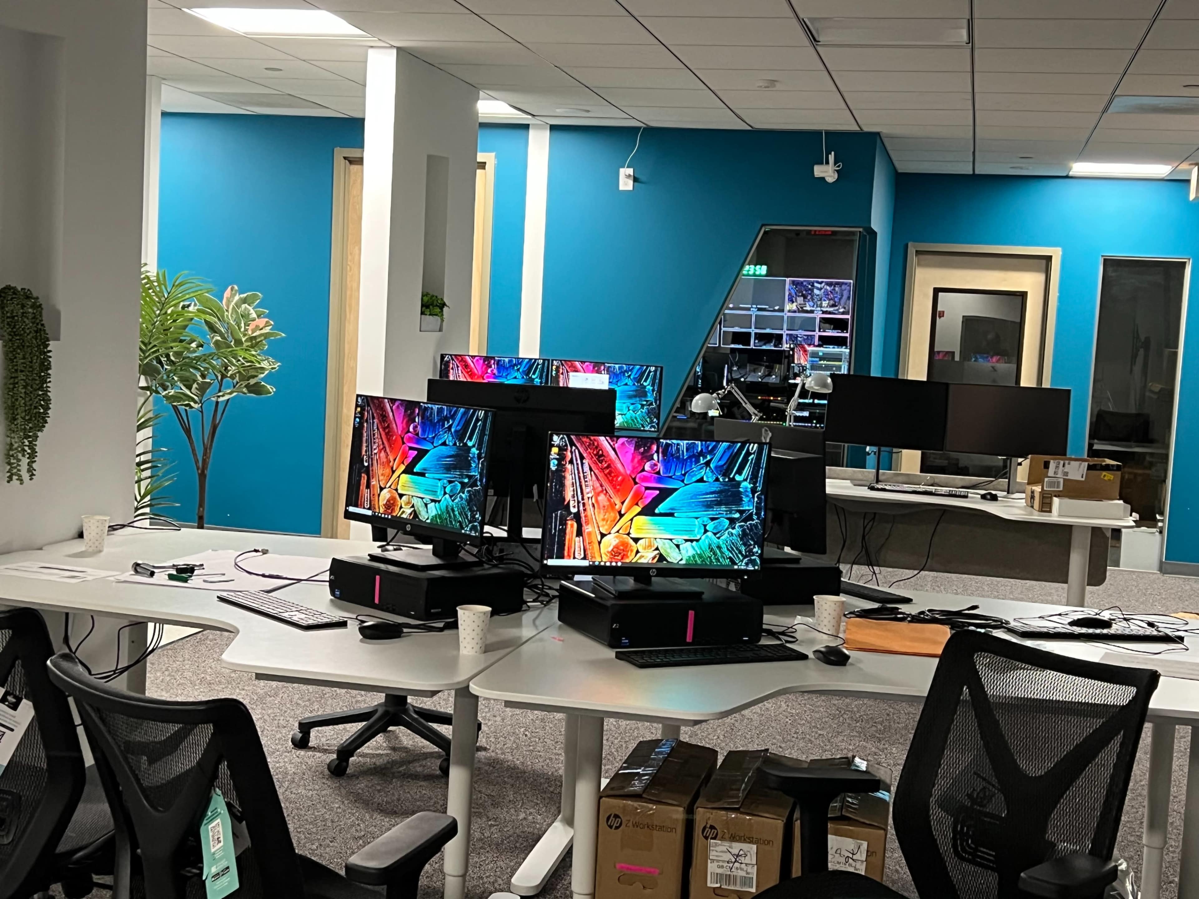 The image shows an office space with multiple workstations equipped with computer monitors, surrounded by blue walls and a few plants.