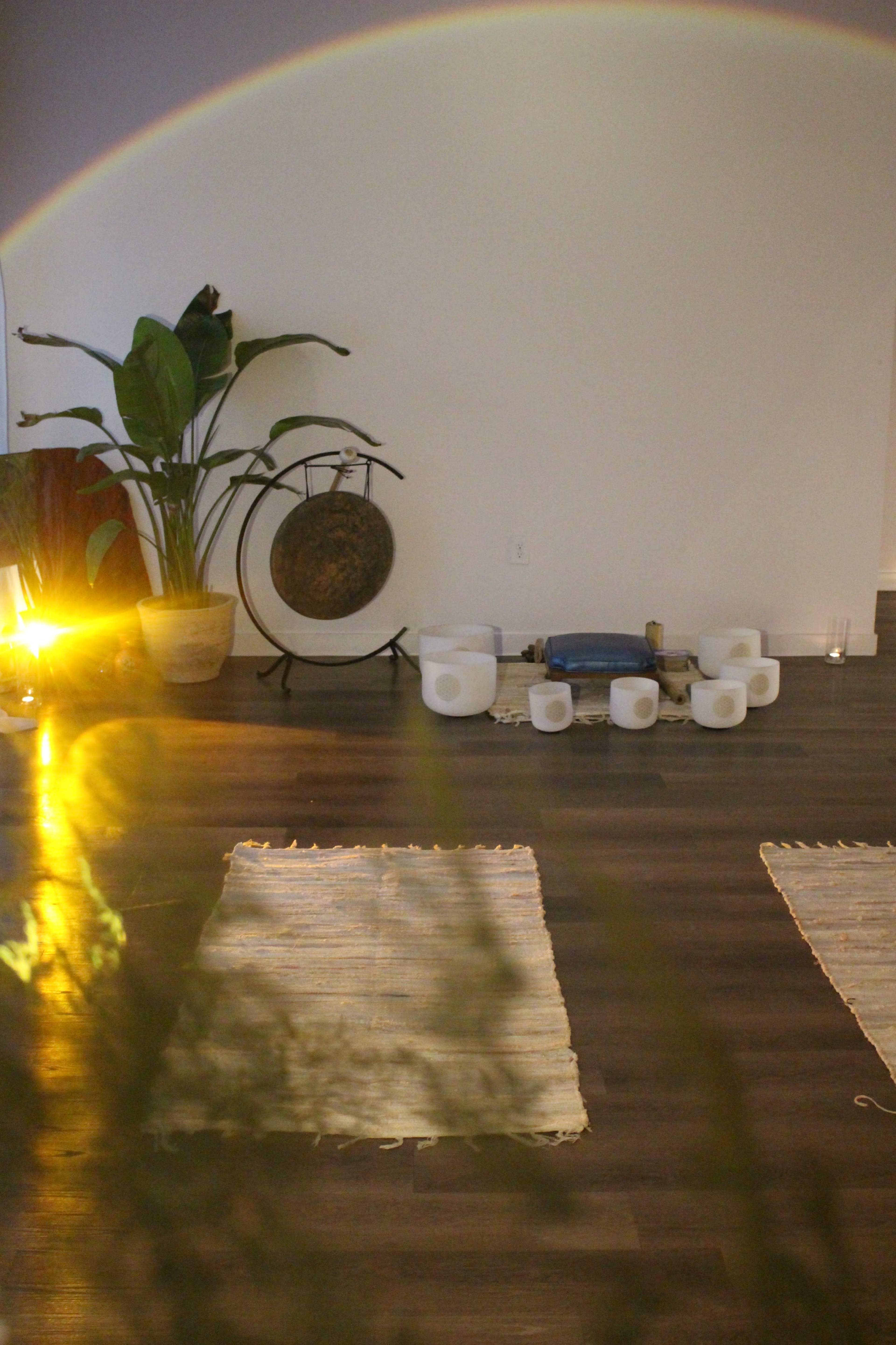 The image shows a tranquil room with woven mats on the floor, a gong, a blue cushion, and several white crystal bowls arranged in a circle.