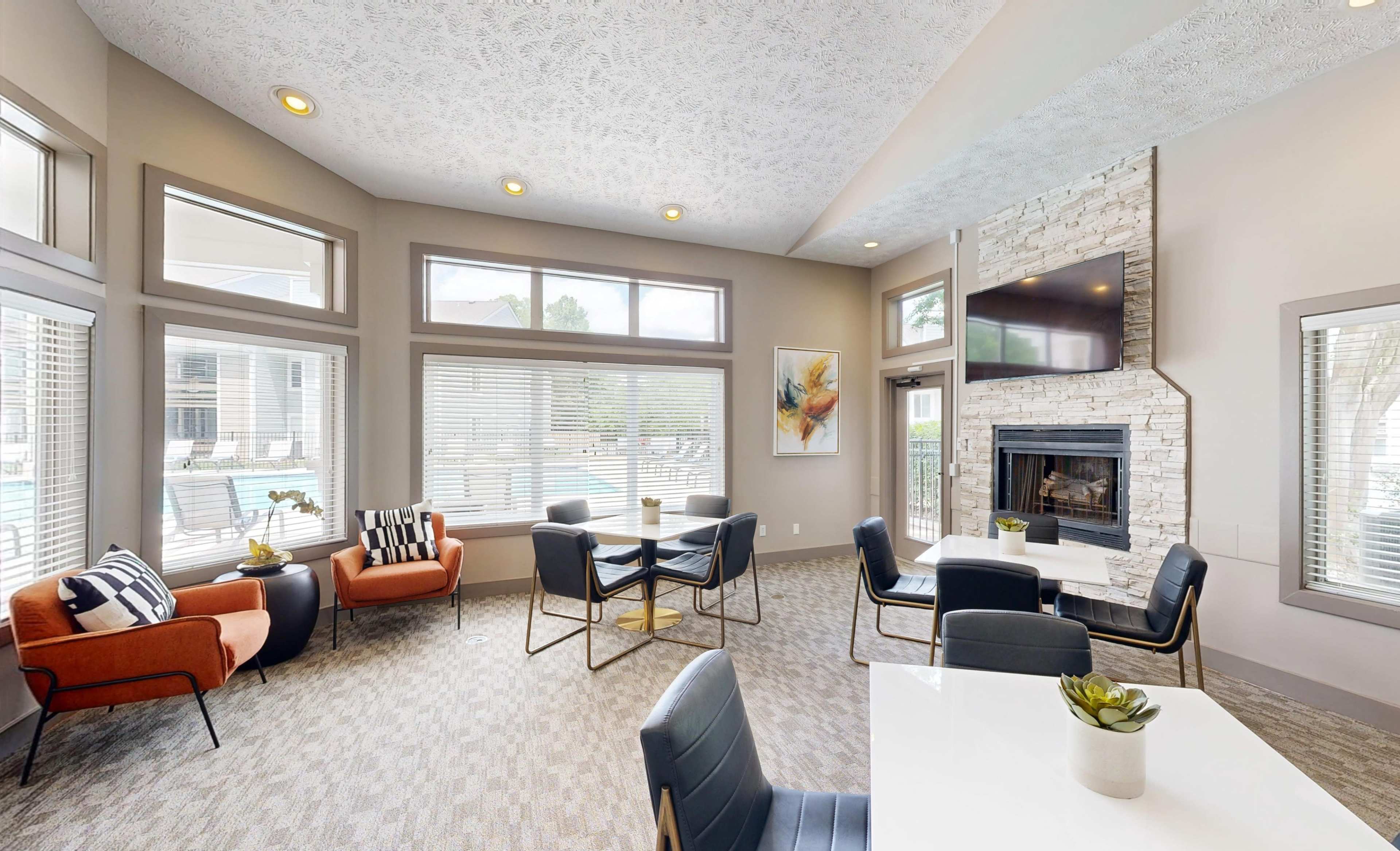 The image shows a cozy community lounge area with seating, a dining table, large windows, and a fireplace, all styled in neutral tones.