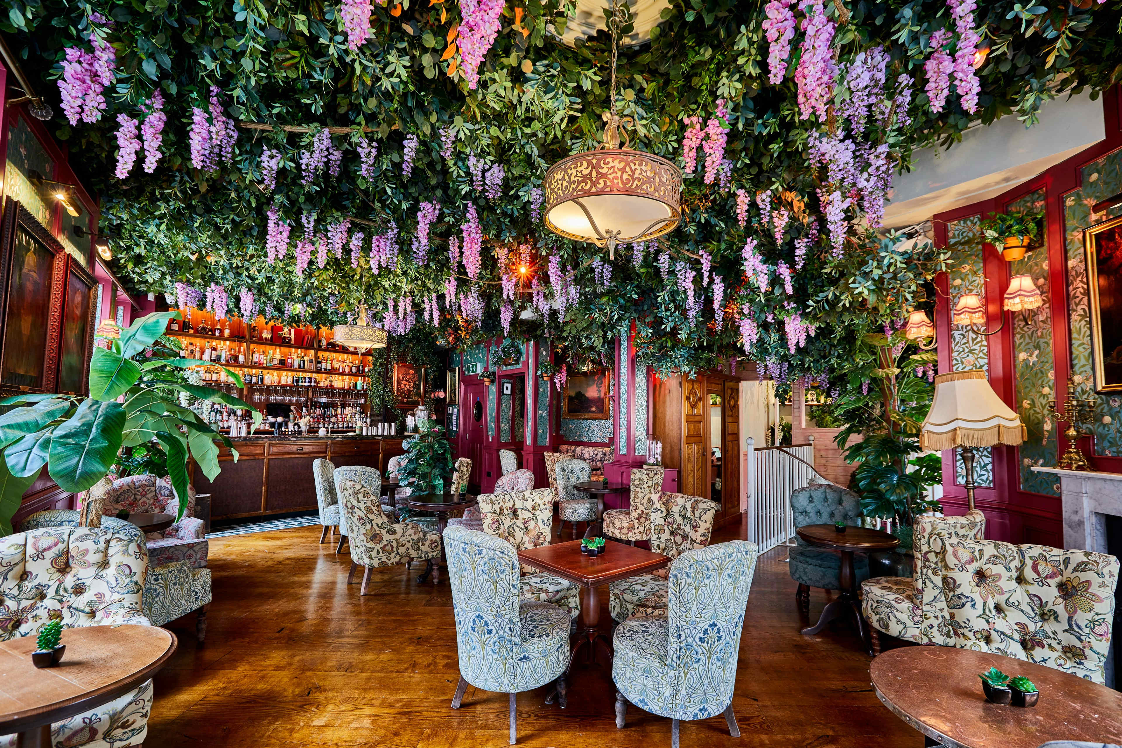 The image features a lushly decorated restaurant interior with floral arrangements hanging from the ceiling and cozy seating areas surrounded by greenery.