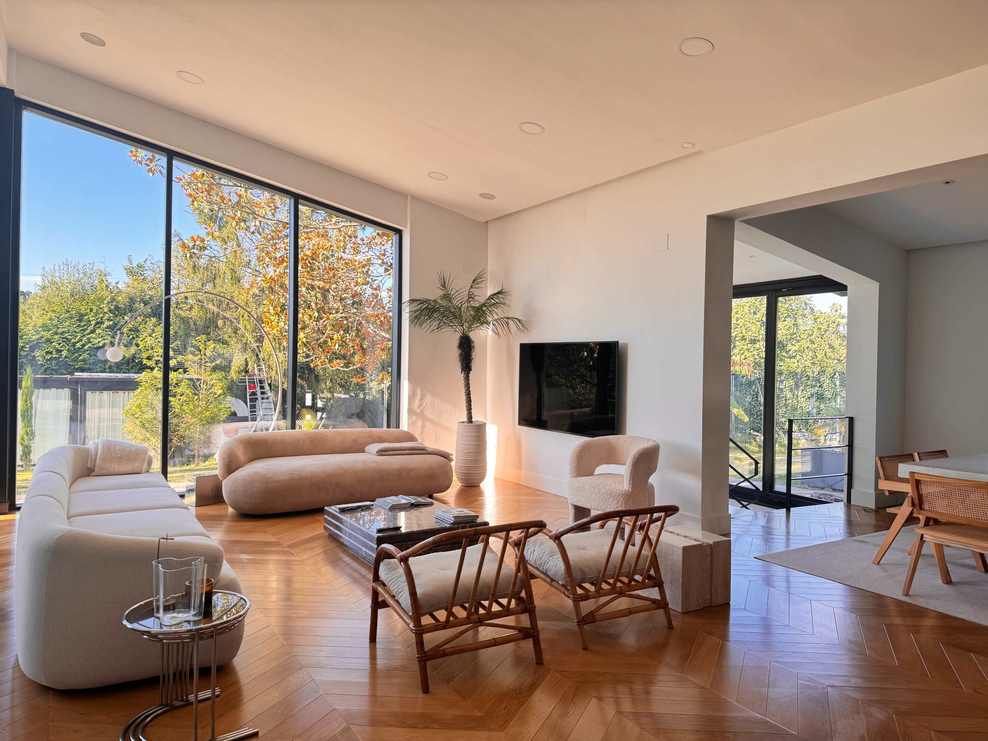 The image shows a modern living room with large windows, a curved sofa, a round coffee table, rattan chairs, and a dining area in the background.