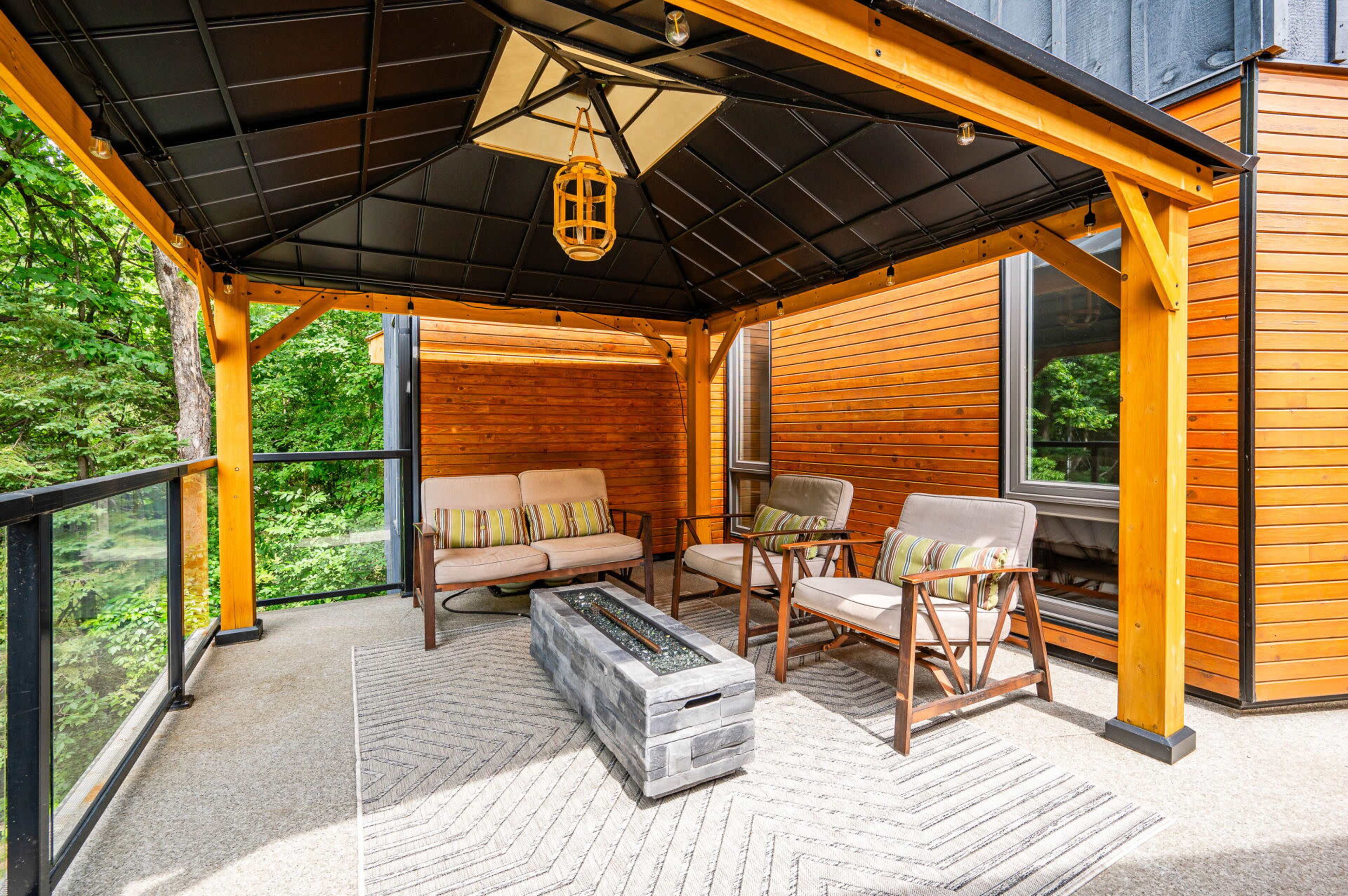 A covered outdoor seating area features chairs and a table with a fire pit, surrounded by greenery.