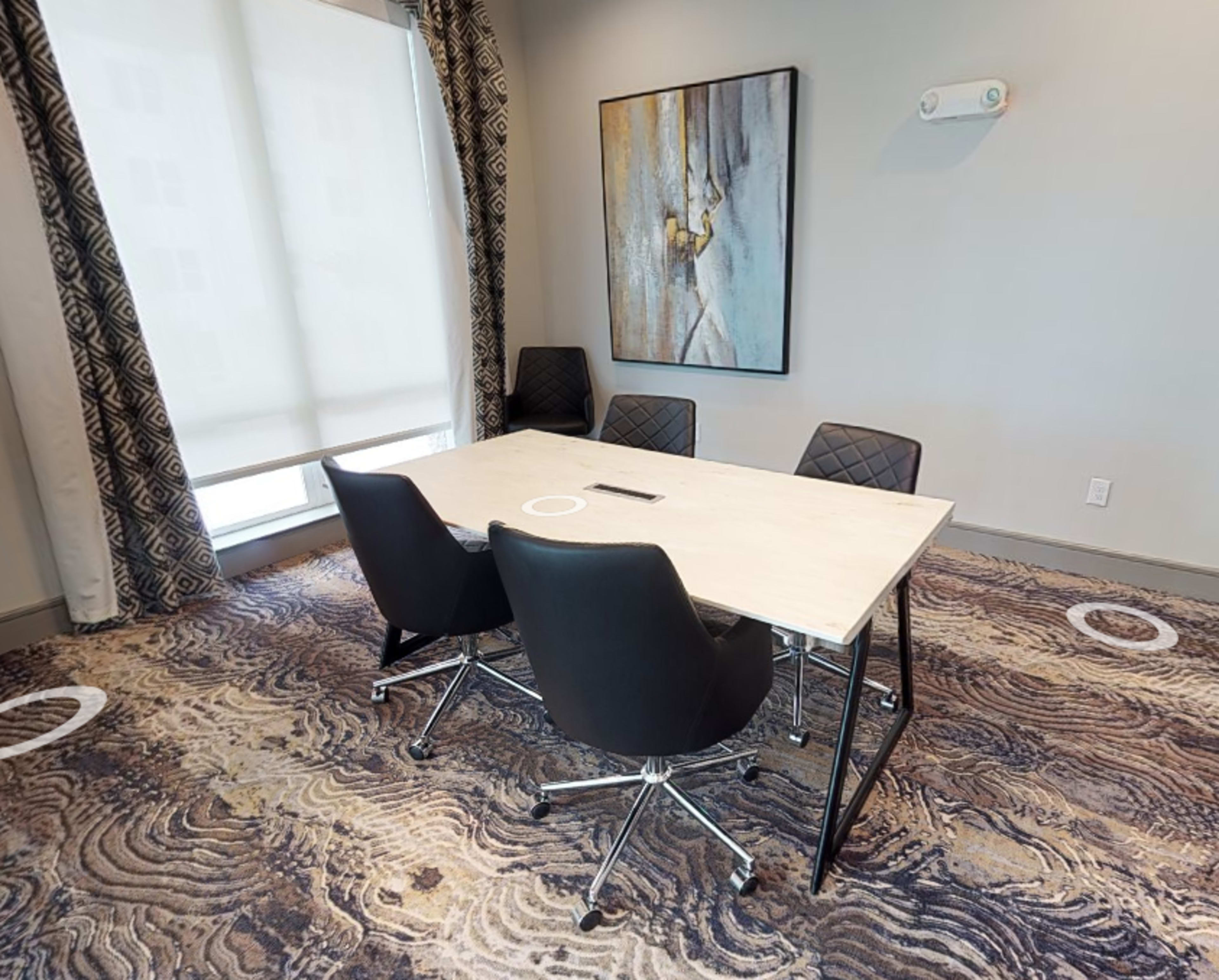 A conference room features a table surrounded by four black chairs, with large windows and abstract artwork on the wall.