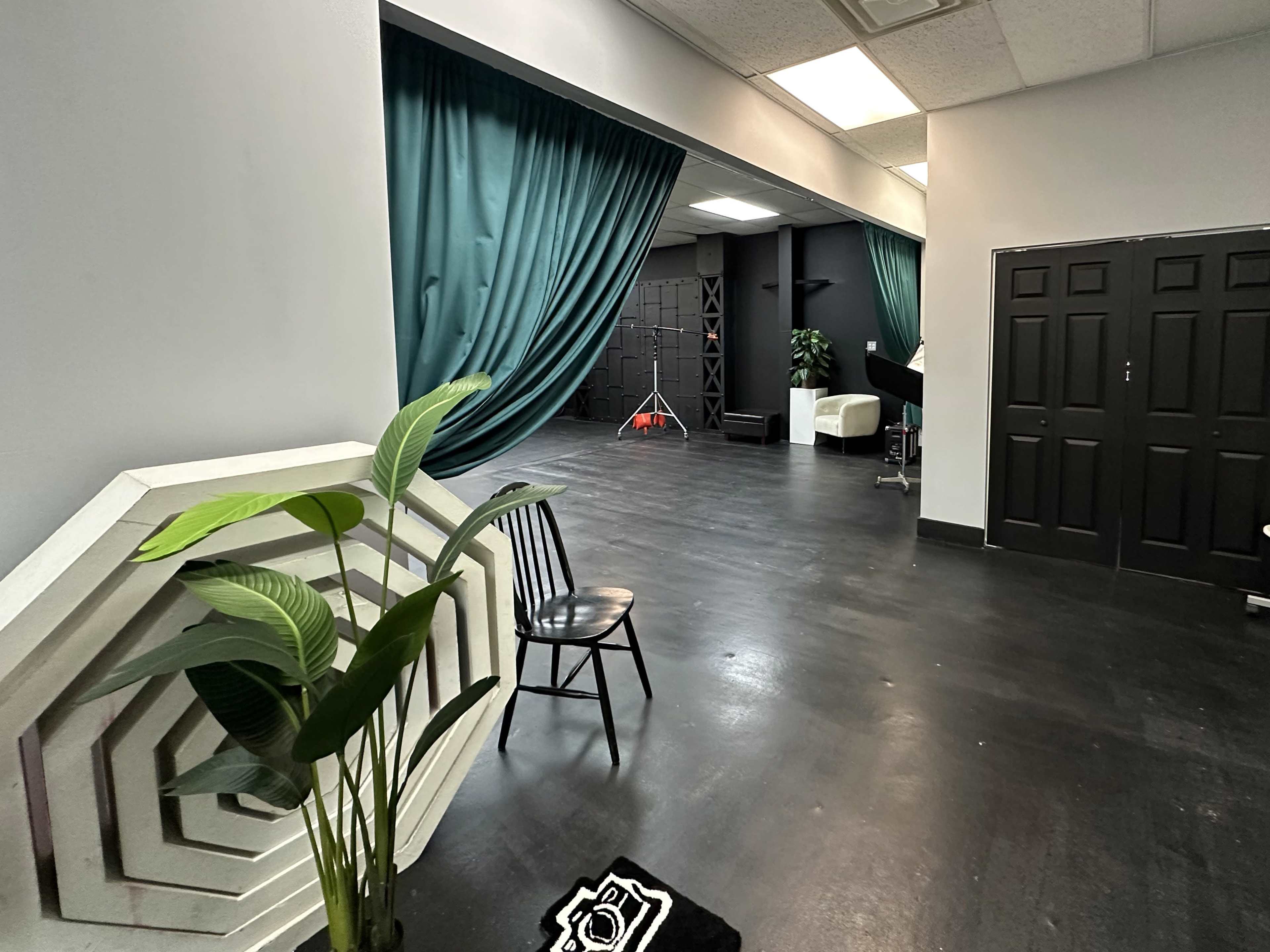An interior space with dark flooring, a chair beside a plant, and curtains dividing areas, along with a studio setup in the background.