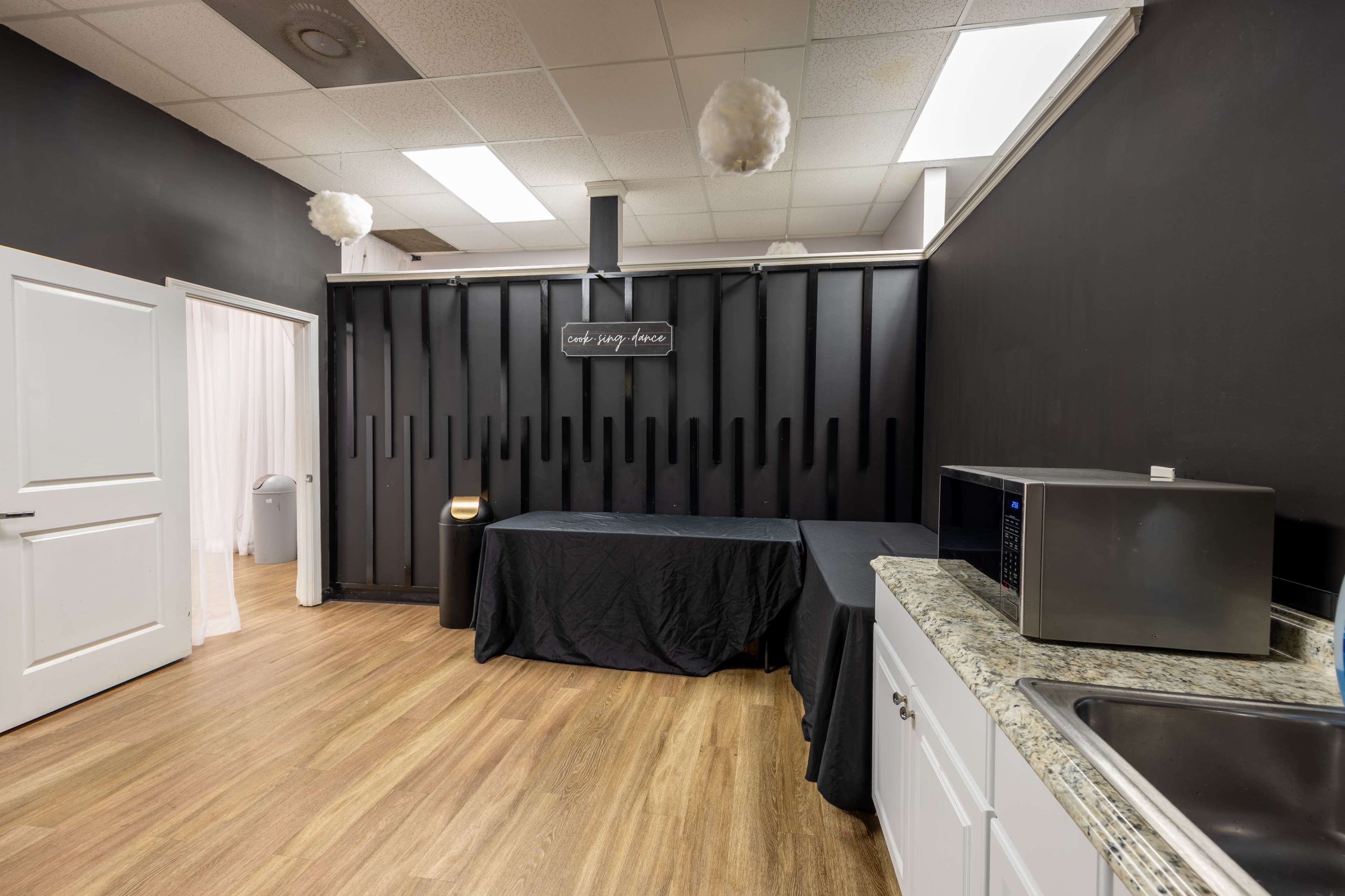 The image shows a room with dark walls, a countertop with a sink and microwave, and two tables covered with black cloth.