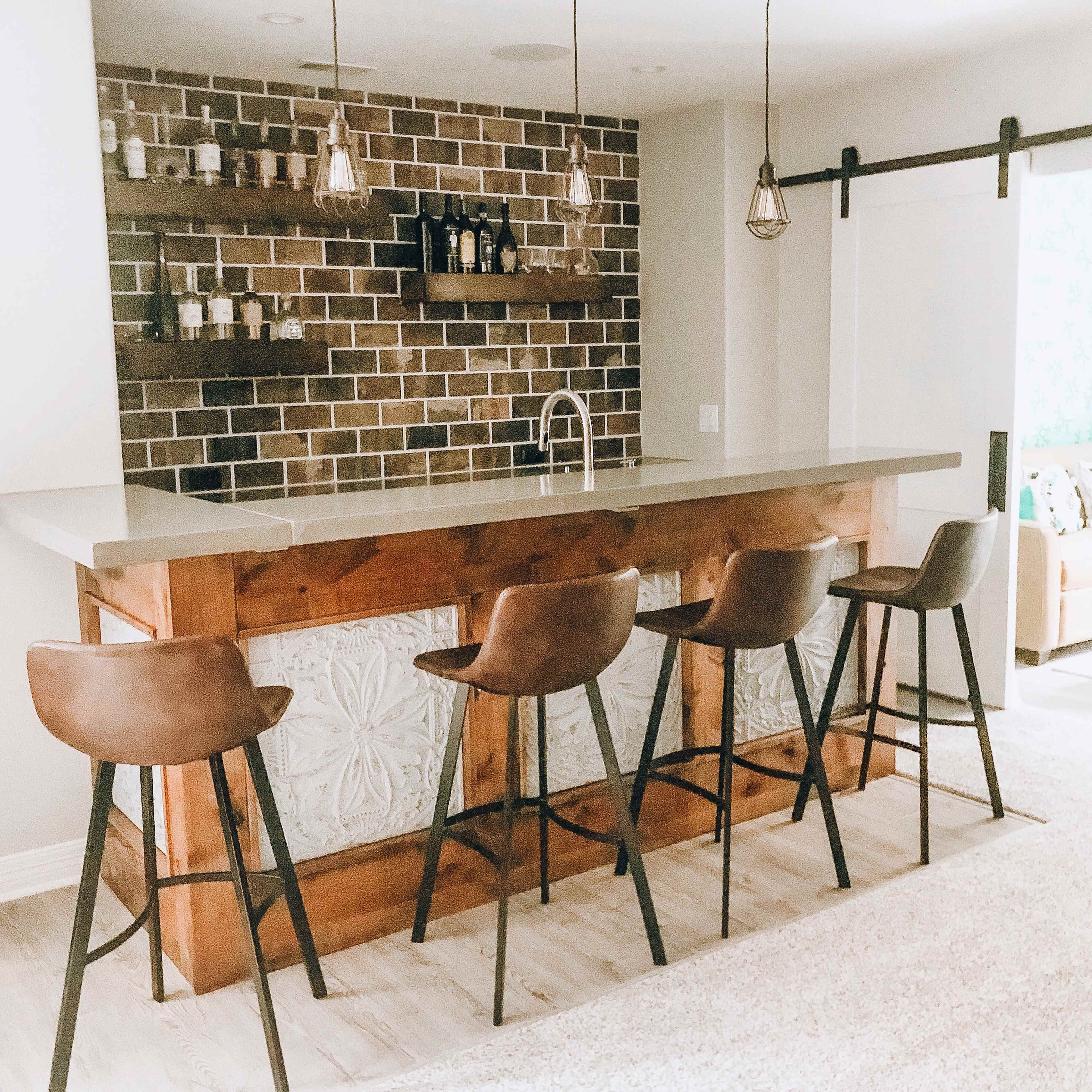 A modern home bar features a wooden counter with four high stools, a backsplash of dark bricks, and shelves displaying bottles and jars.