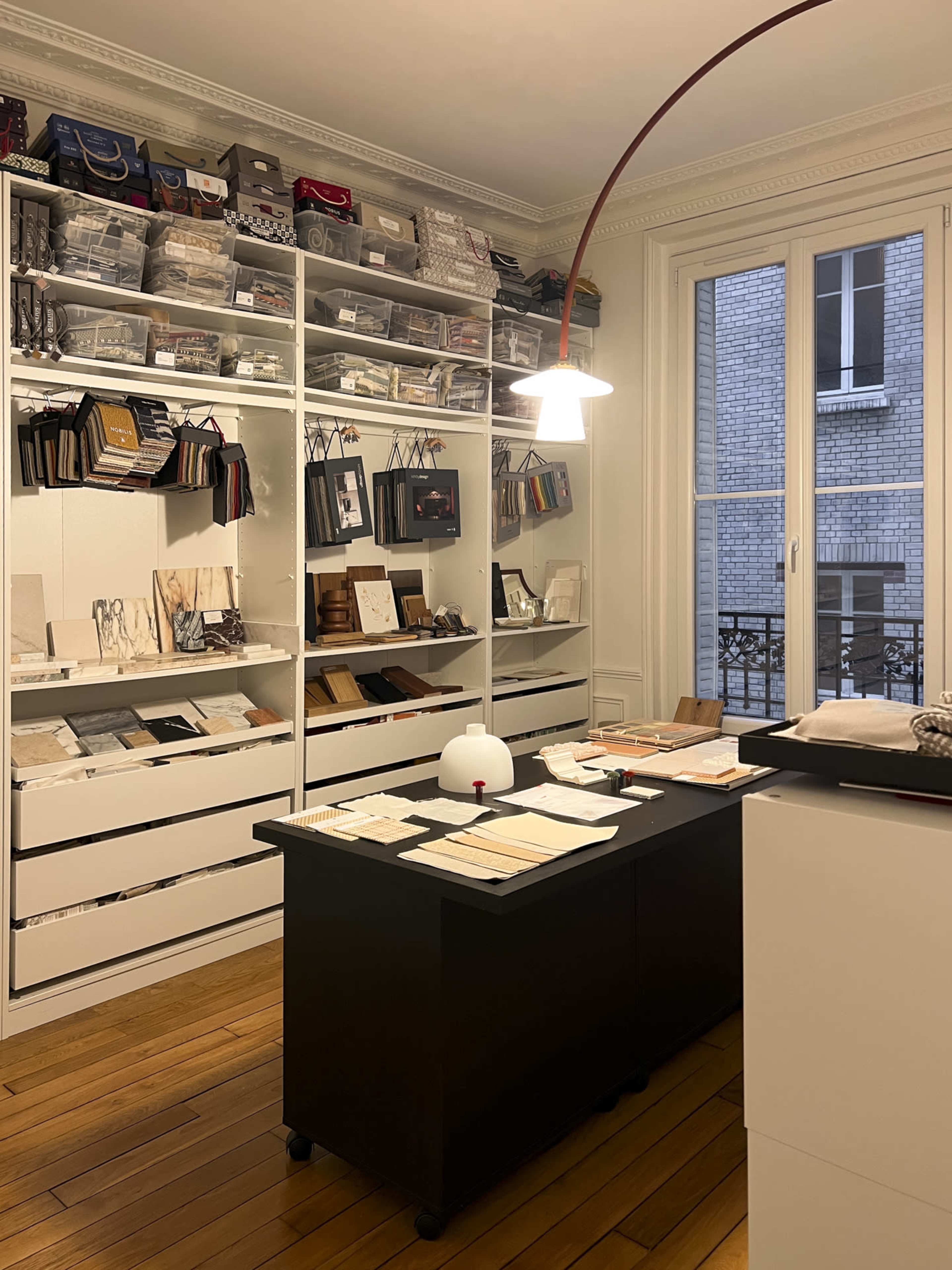 The image shows a well-organized workspace featuring shelves filled with materials and samples, a central table covered with design items, and a large window allowing natural light to enter the room.