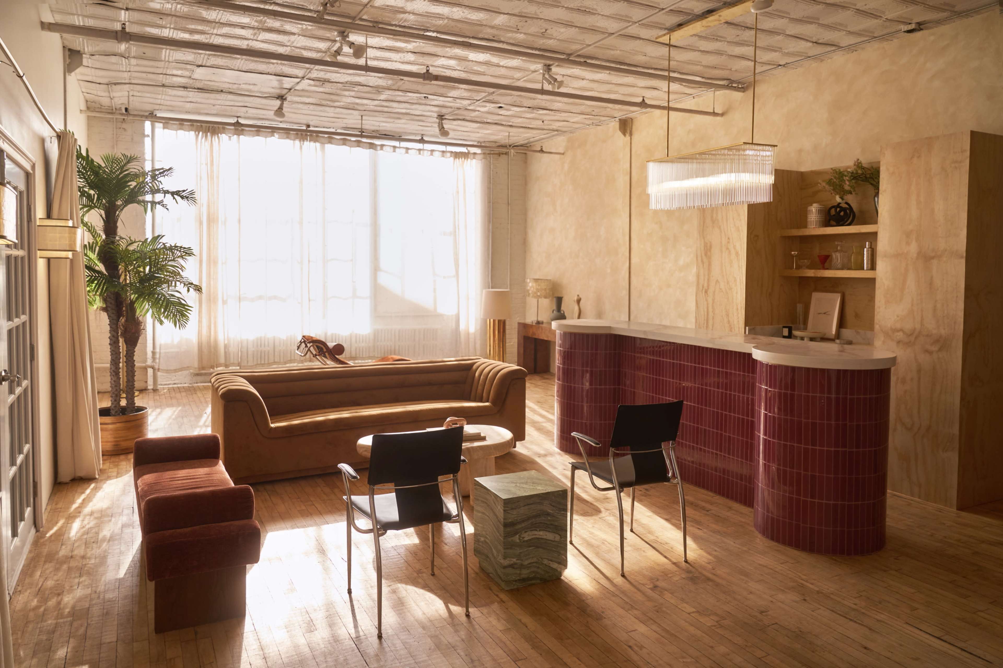 The image shows a spacious, well-lit room featuring a brown sofa, two black chairs, a round coffee table, and a bar-style counter, with large windows allowing ample natural light.