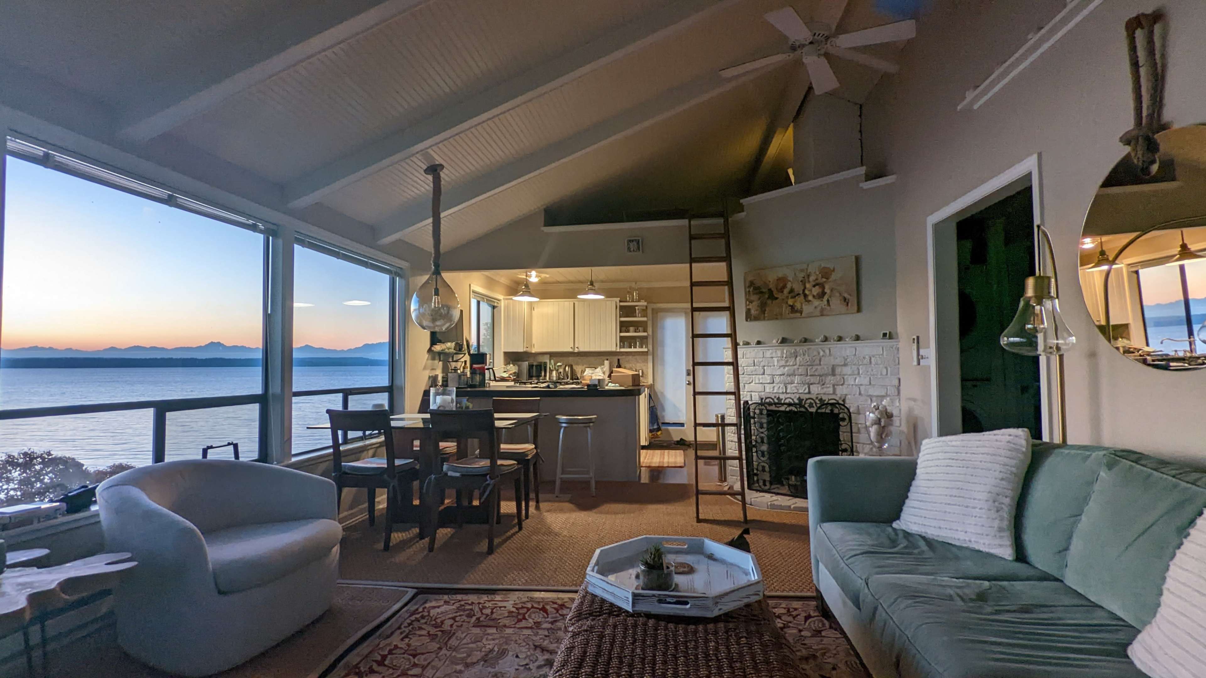 A cozy coastal living room with a view of the ocean at sunset, featuring a light-colored sofa, a dining table, and a modern kitchen in the background.