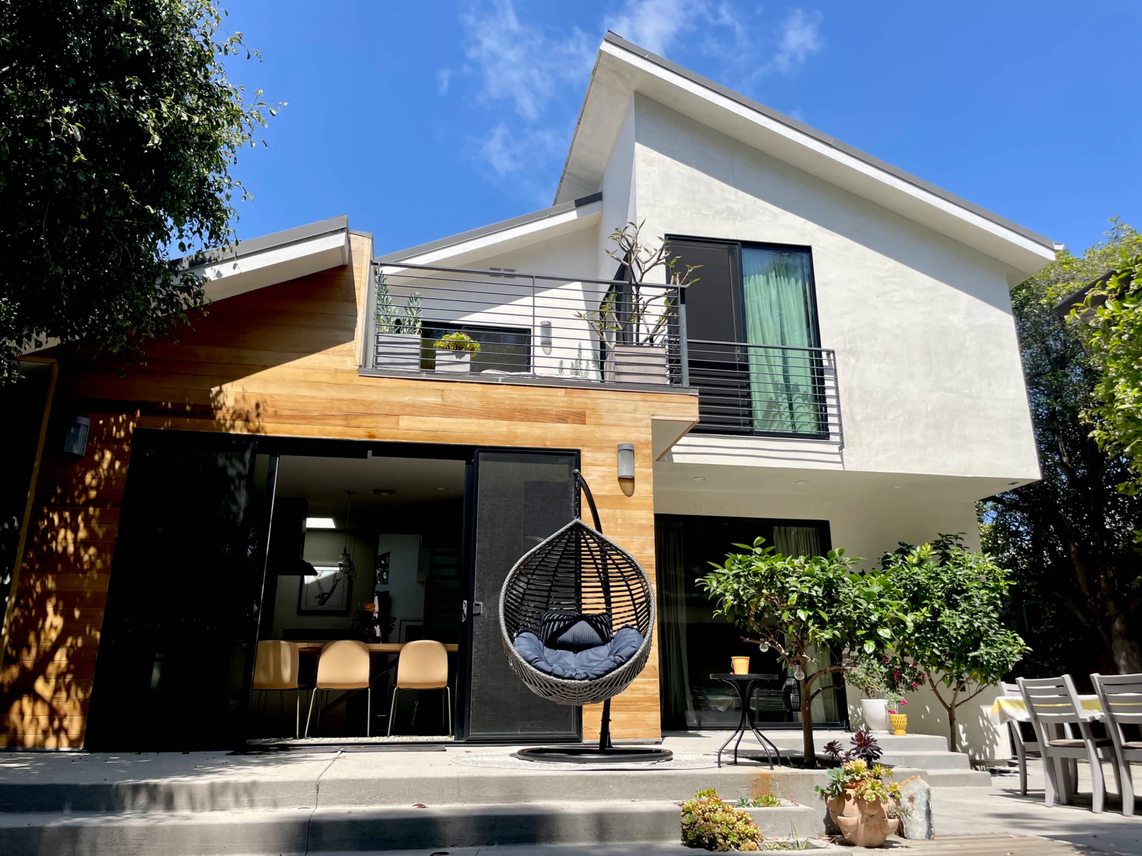 A modern two-story house features a combination of wood and white exterior, with a hanging chair on the patio and large windows.