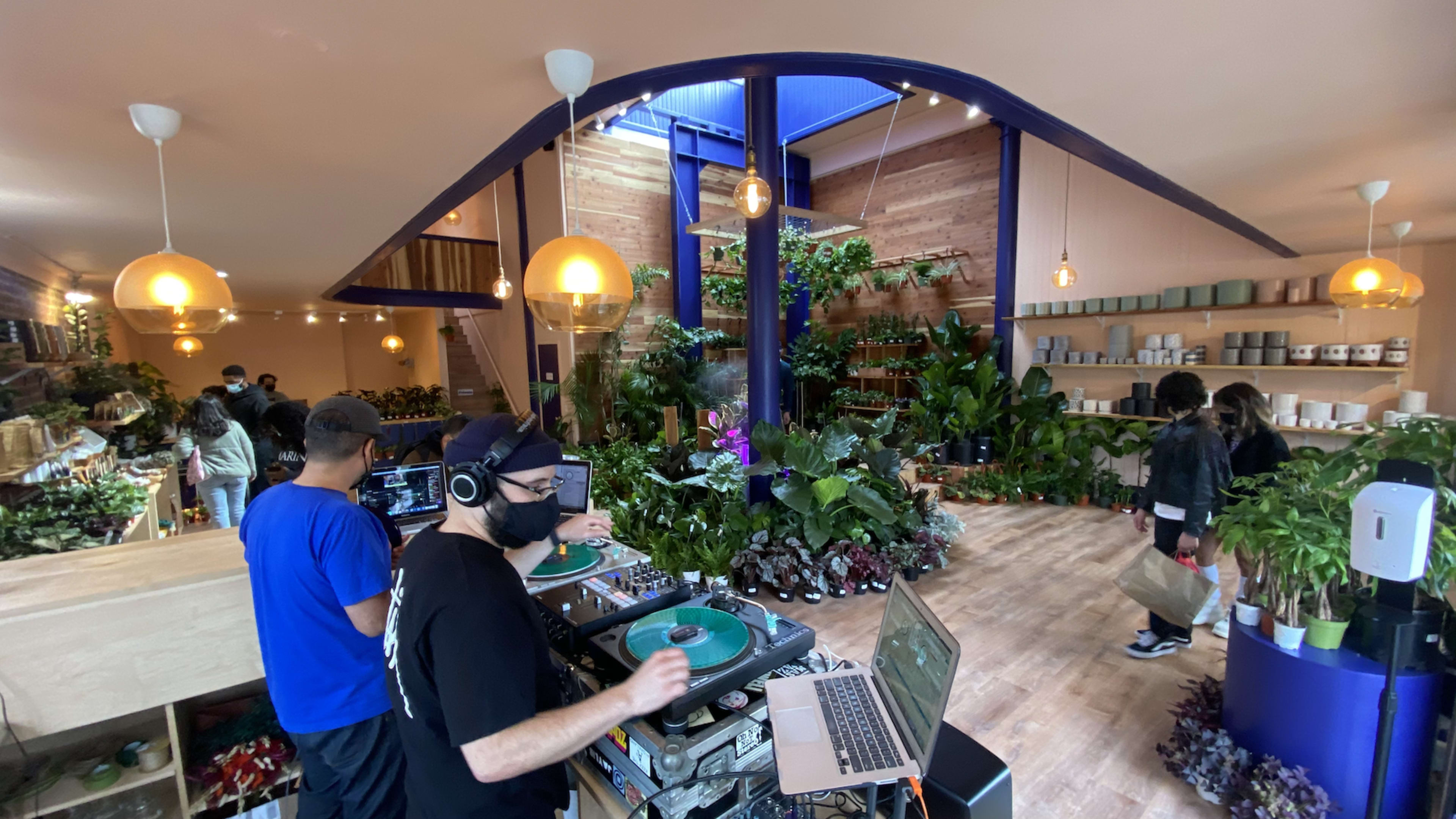 A DJ stands behind turntables in a plant-filled store while customers browse the greenery.