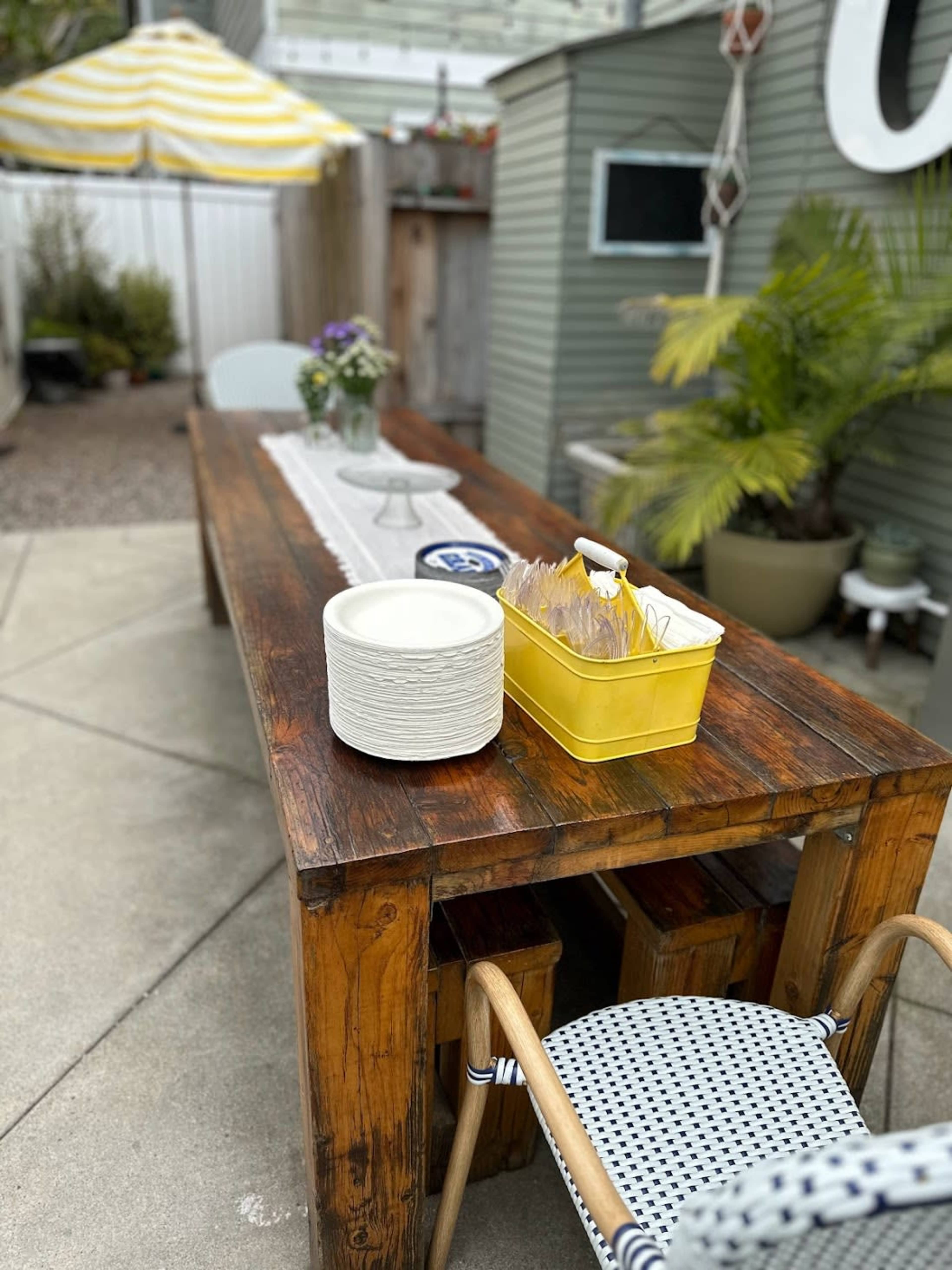A wooden outdoor dining table is set with a white bowl, a yellow container holding utensils, and surrounded by chairs in a landscaped patio area.