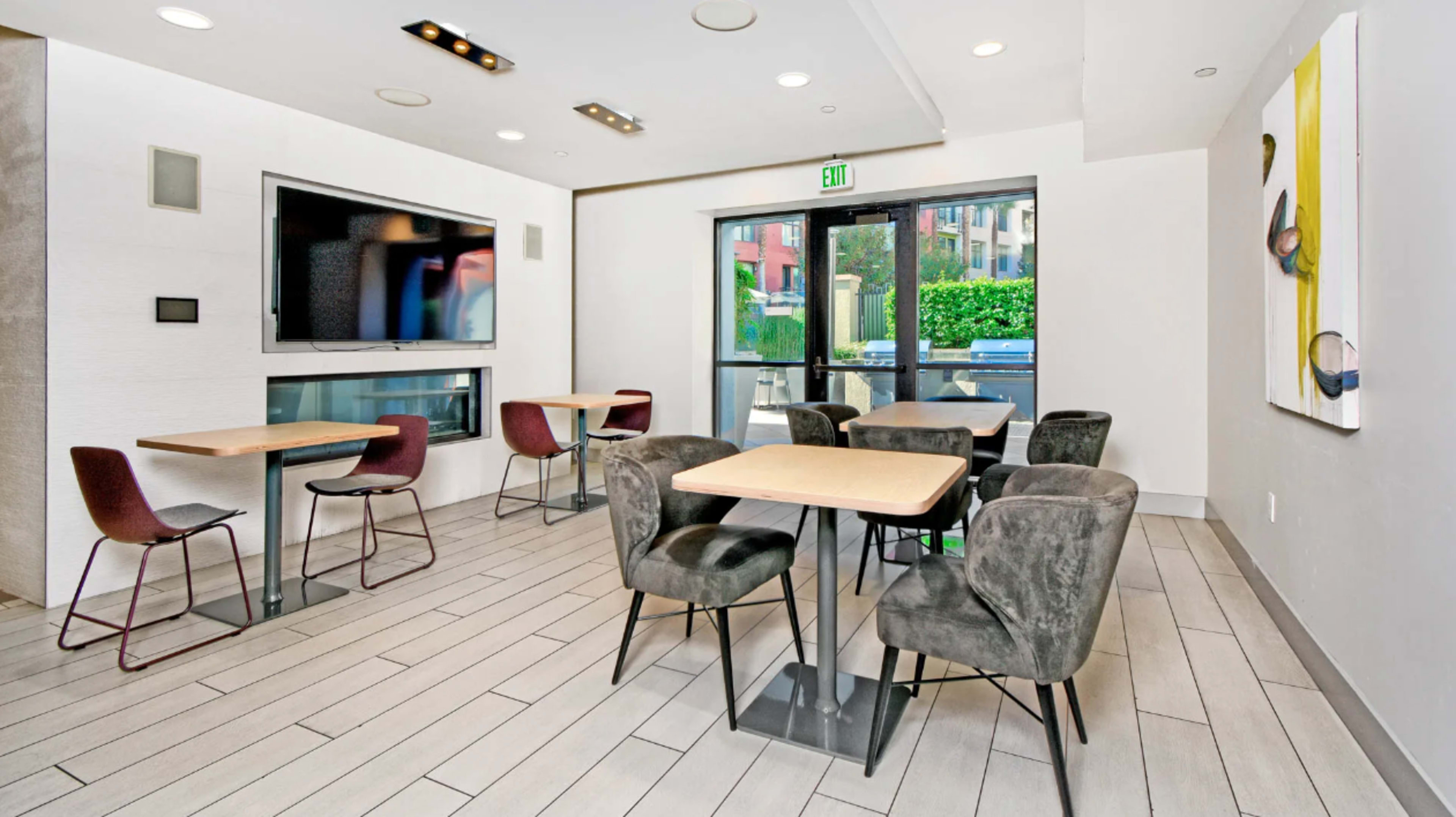 A modern interior space with several tables and chairs, a large wall-mounted television, and a glass door leading to an outdoor area.
