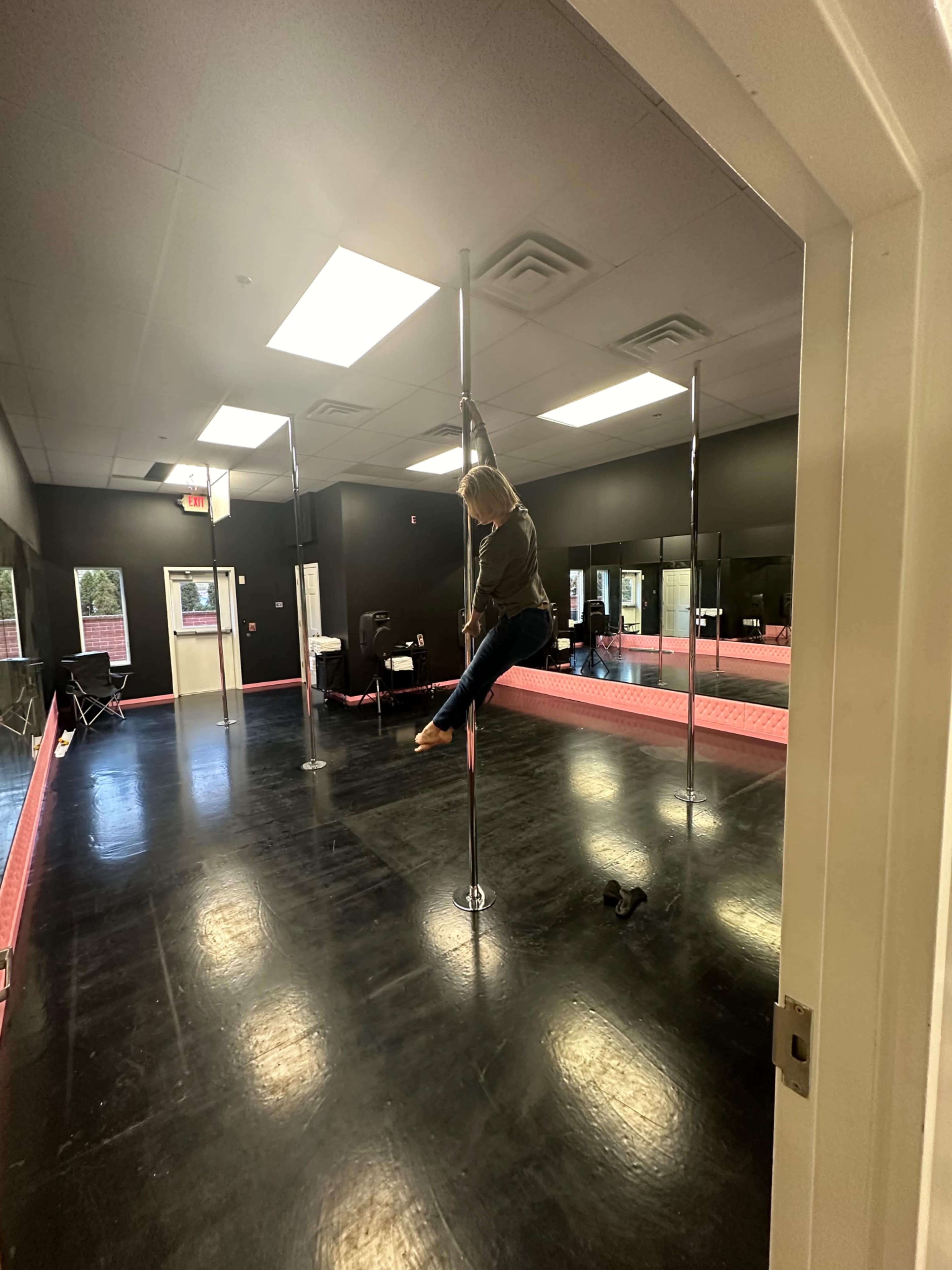 A person is performing a move on a pole in a dance studio with black walls and a shiny floor.