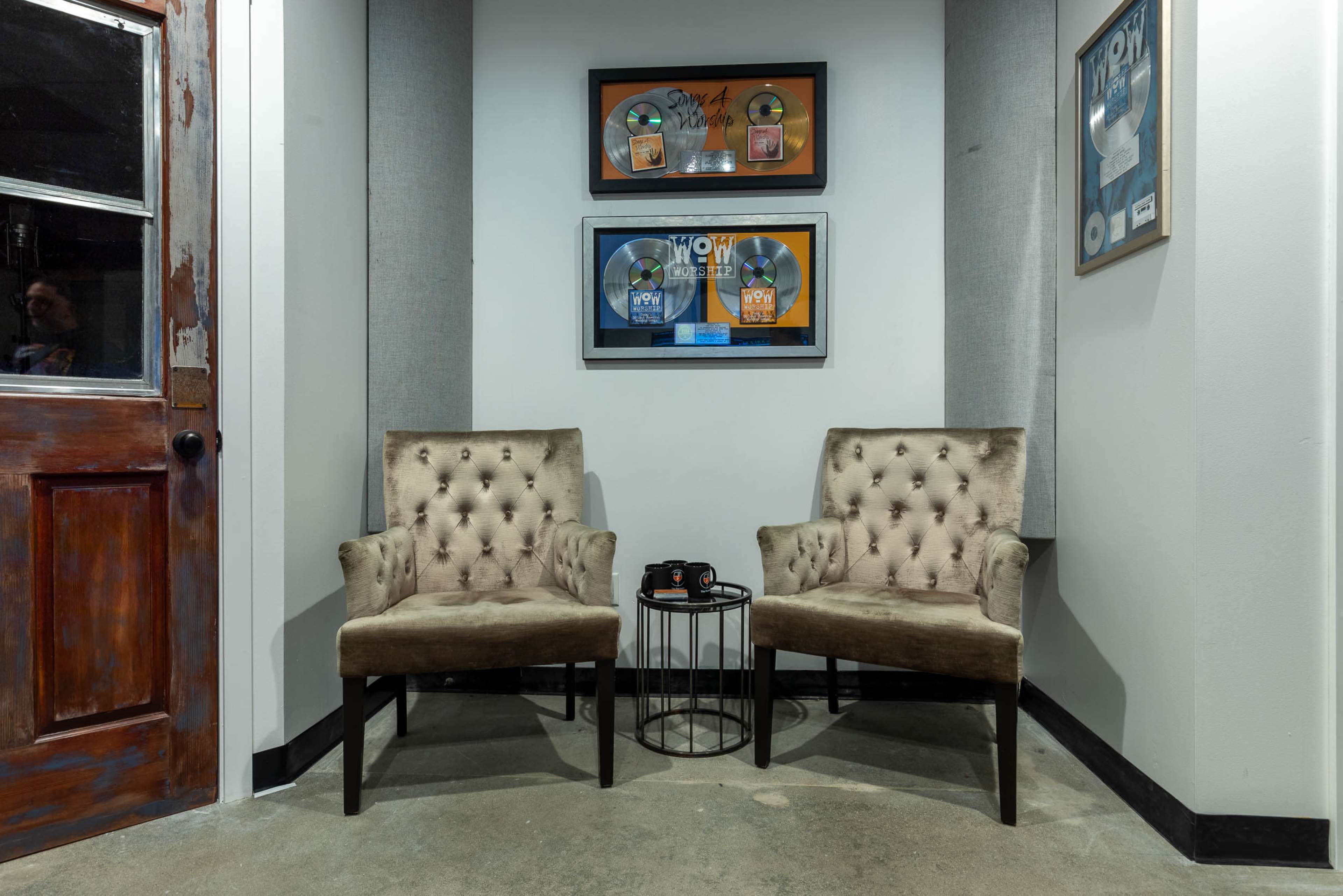 Two tufted armchairs facing a small table in a corner adorned with framed music awards on the wall.