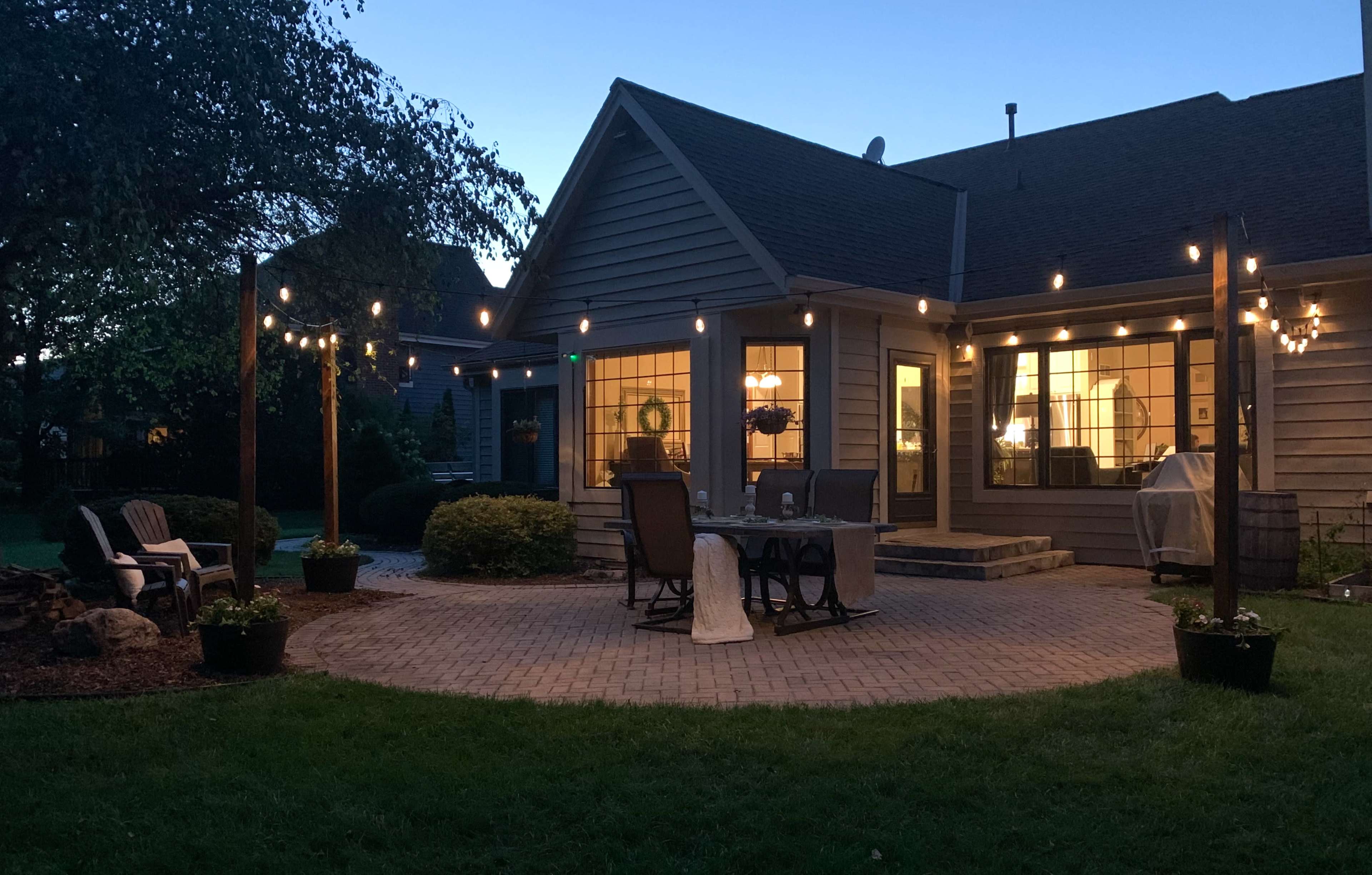 A patio is set up behind a house, illuminated by string lights, with outdoor furniture arranged on a brick surface.