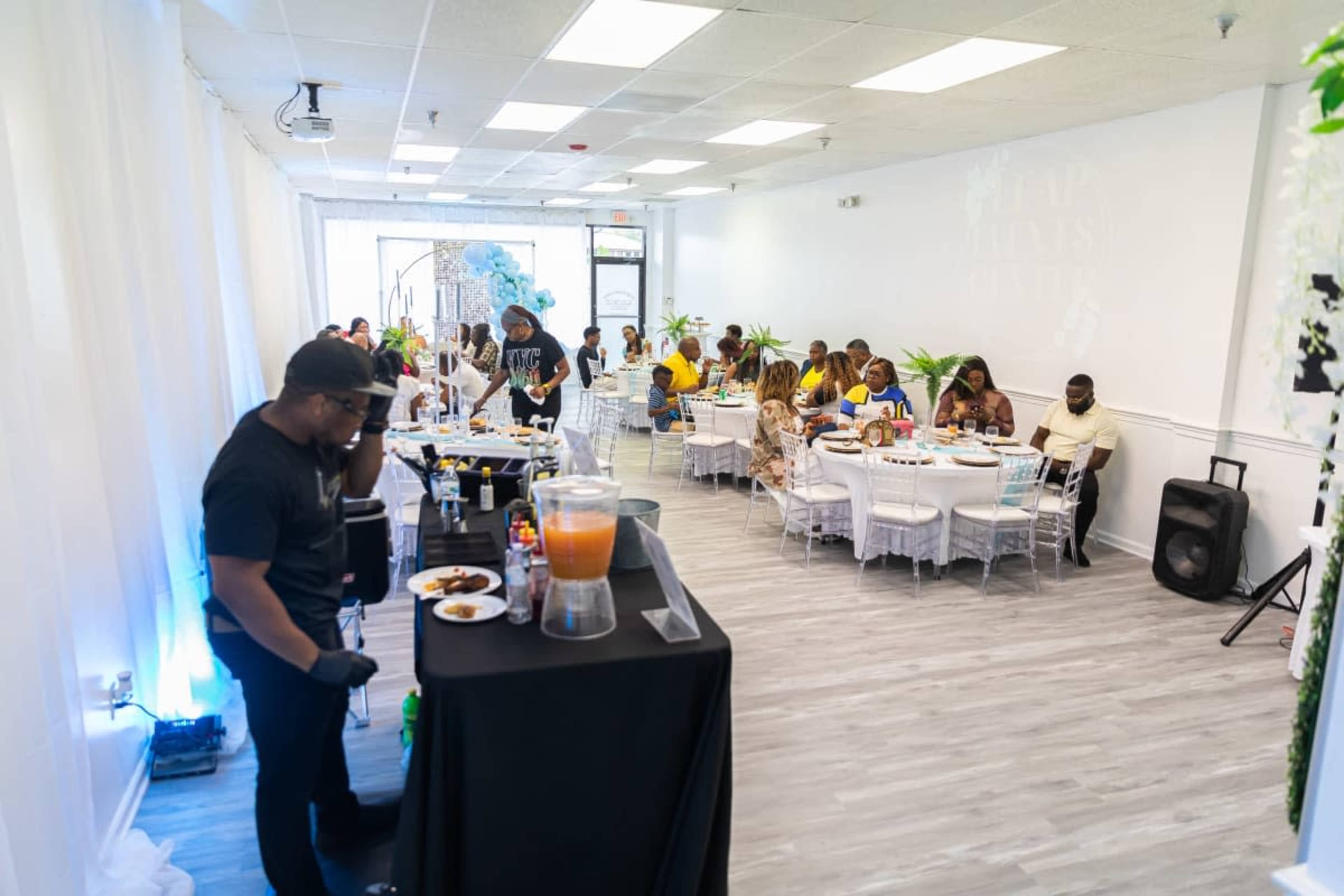 A spacious event room is set up with tables filled with guests enjoying a meal, while a person at a buffet table prepares food.