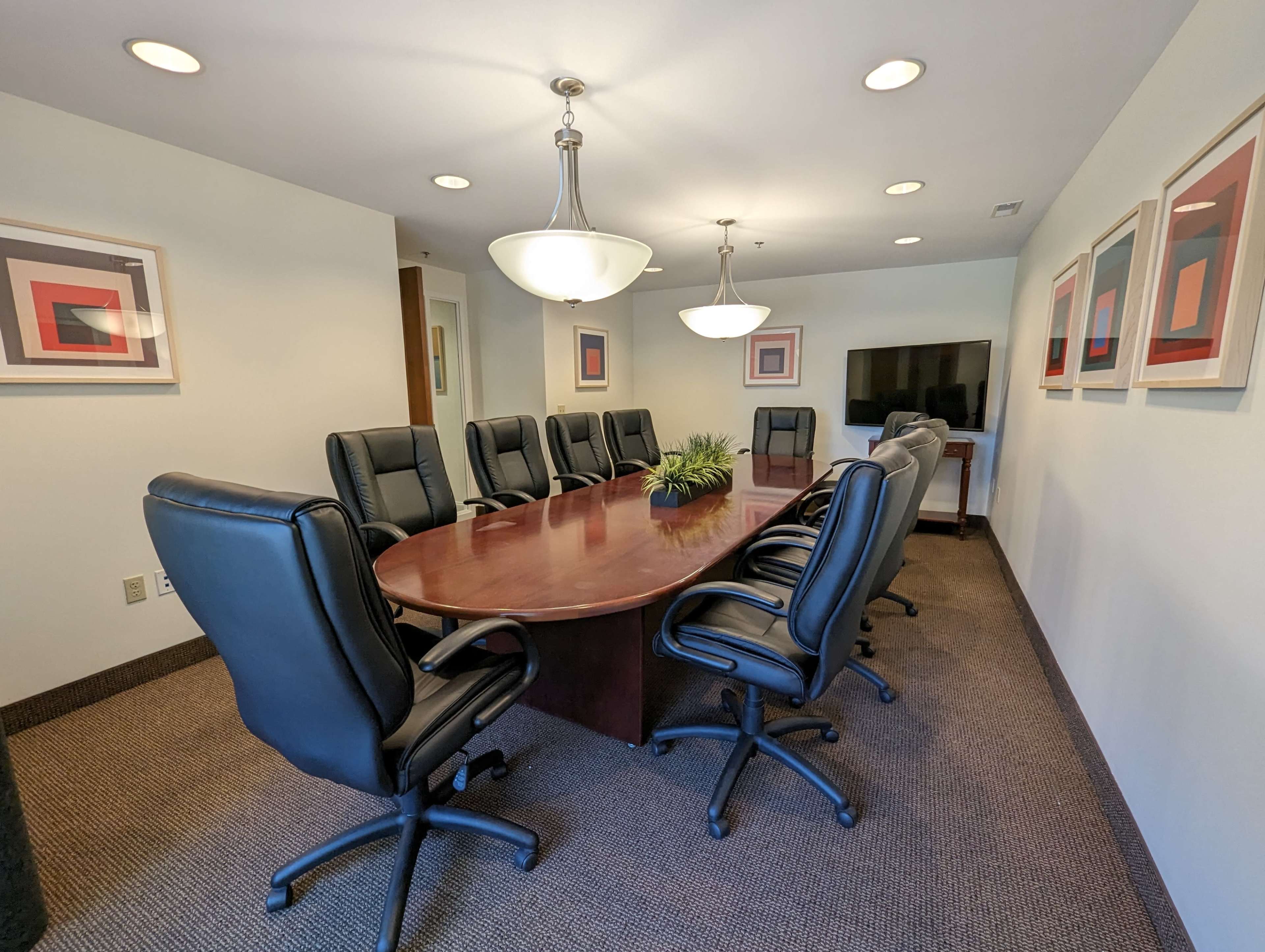 A formal meeting room features a large oval table surrounded by eight black leather chairs, with two additional chairs at one end and modern artwork on the walls.