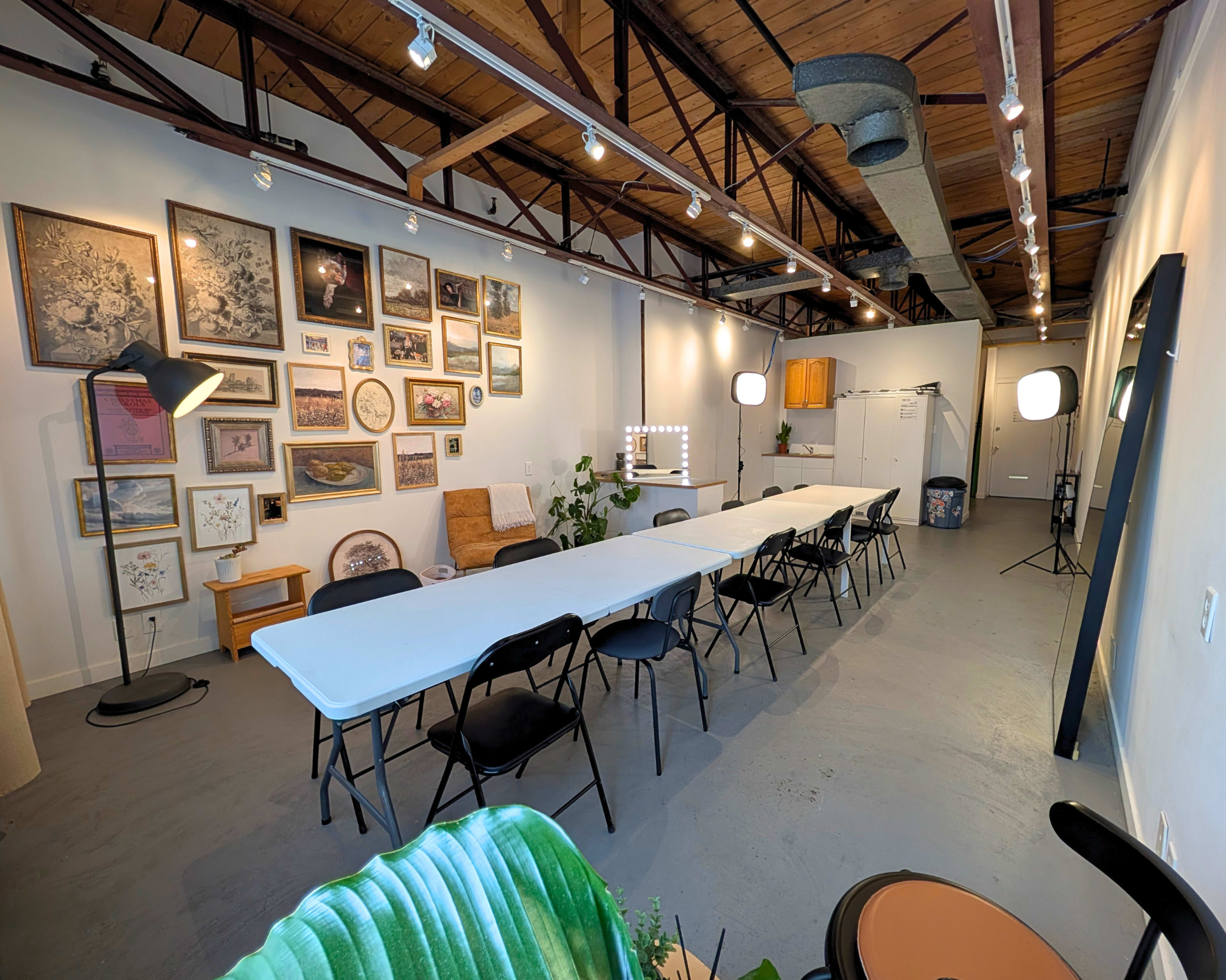 The image shows a spacious room with a long white table set up for a meeting or event, surrounded by black chairs and adorned with framed artwork on the wall.