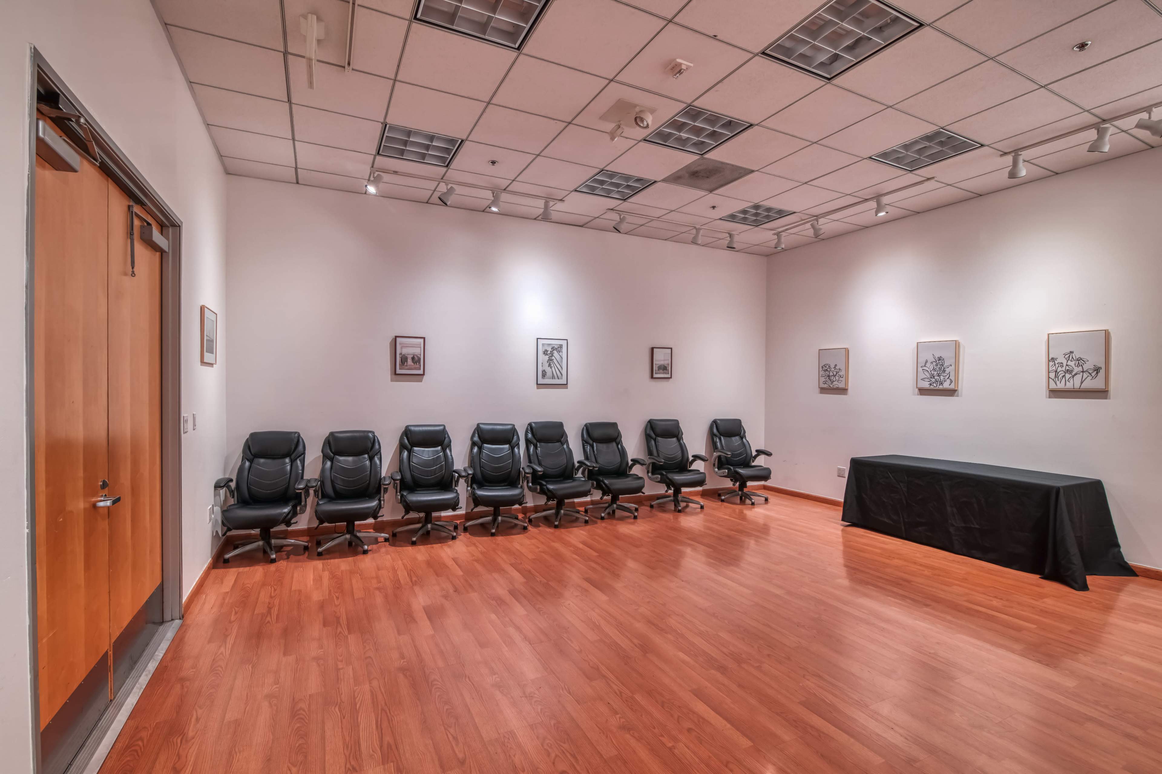 The image shows a sparsely furnished conference room with eight black leather chairs arranged in a row along the wall and a long table covered with a black cloth against another wall.