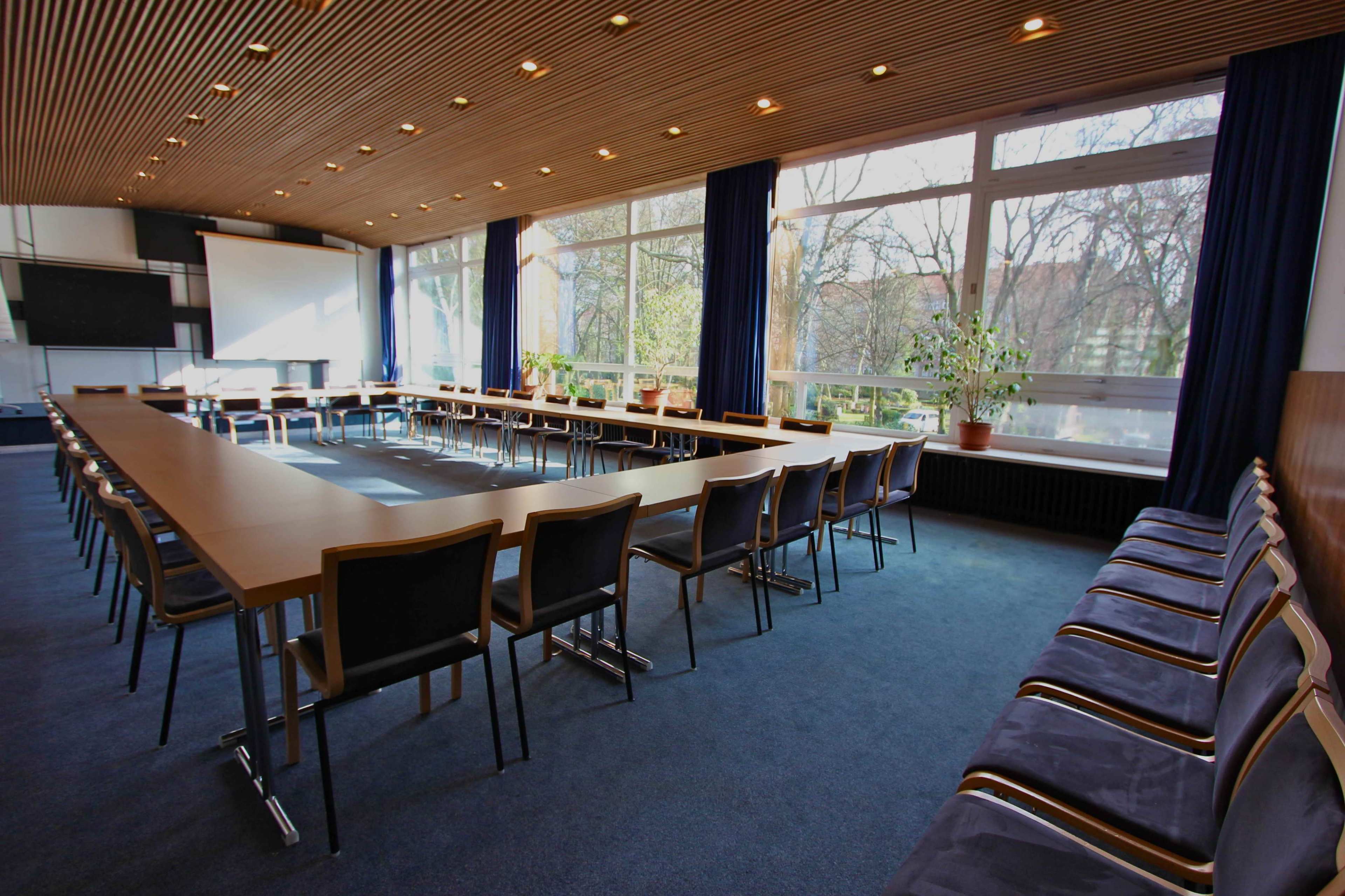 A large conference room features a long rectangular table surrounded by chairs, with large windows letting in natural light and views of trees outside.