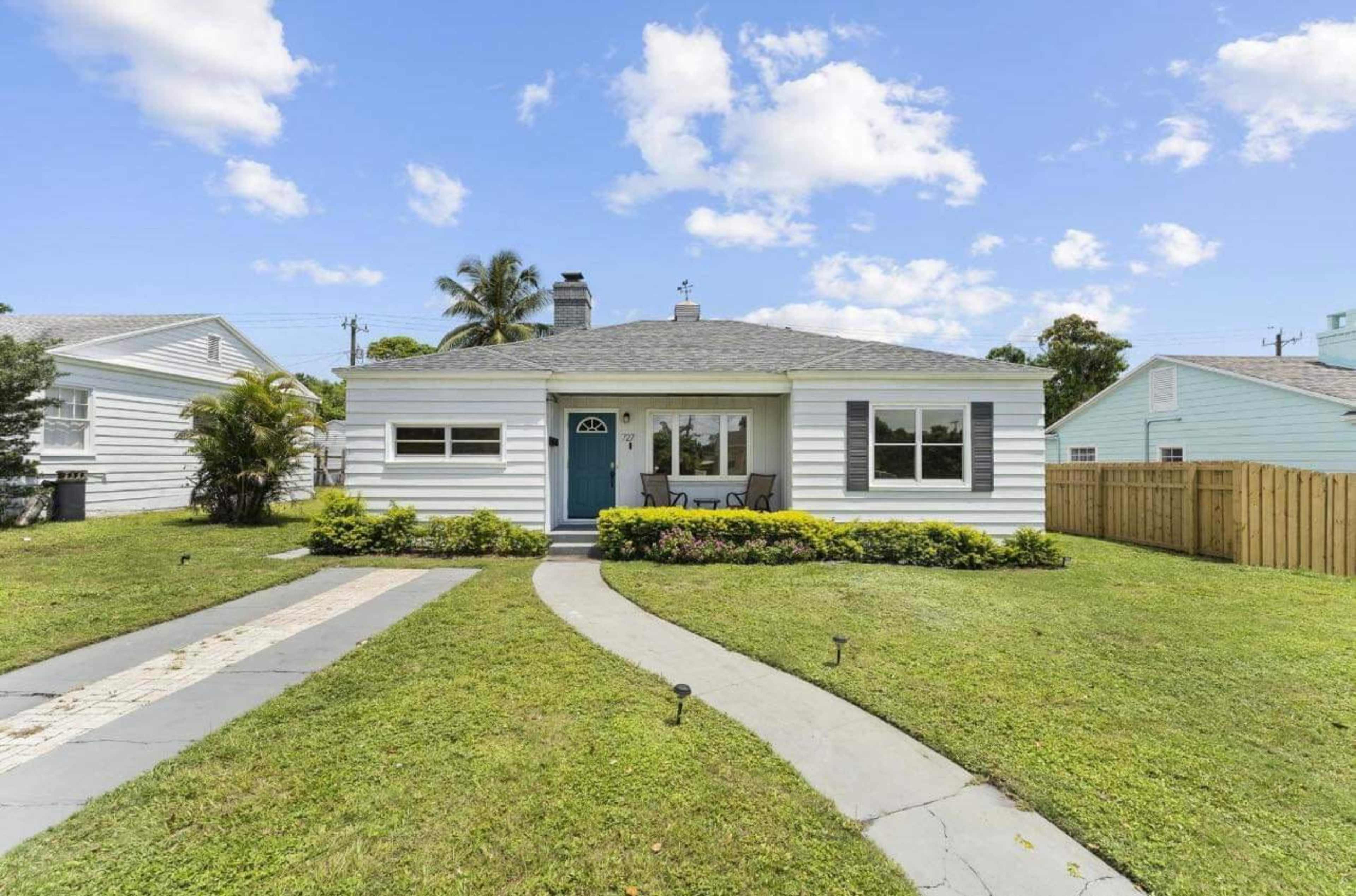 A single-story house with a blue front door sits on a grassy lawn, surrounded by a concrete walkway and low shrubbery.