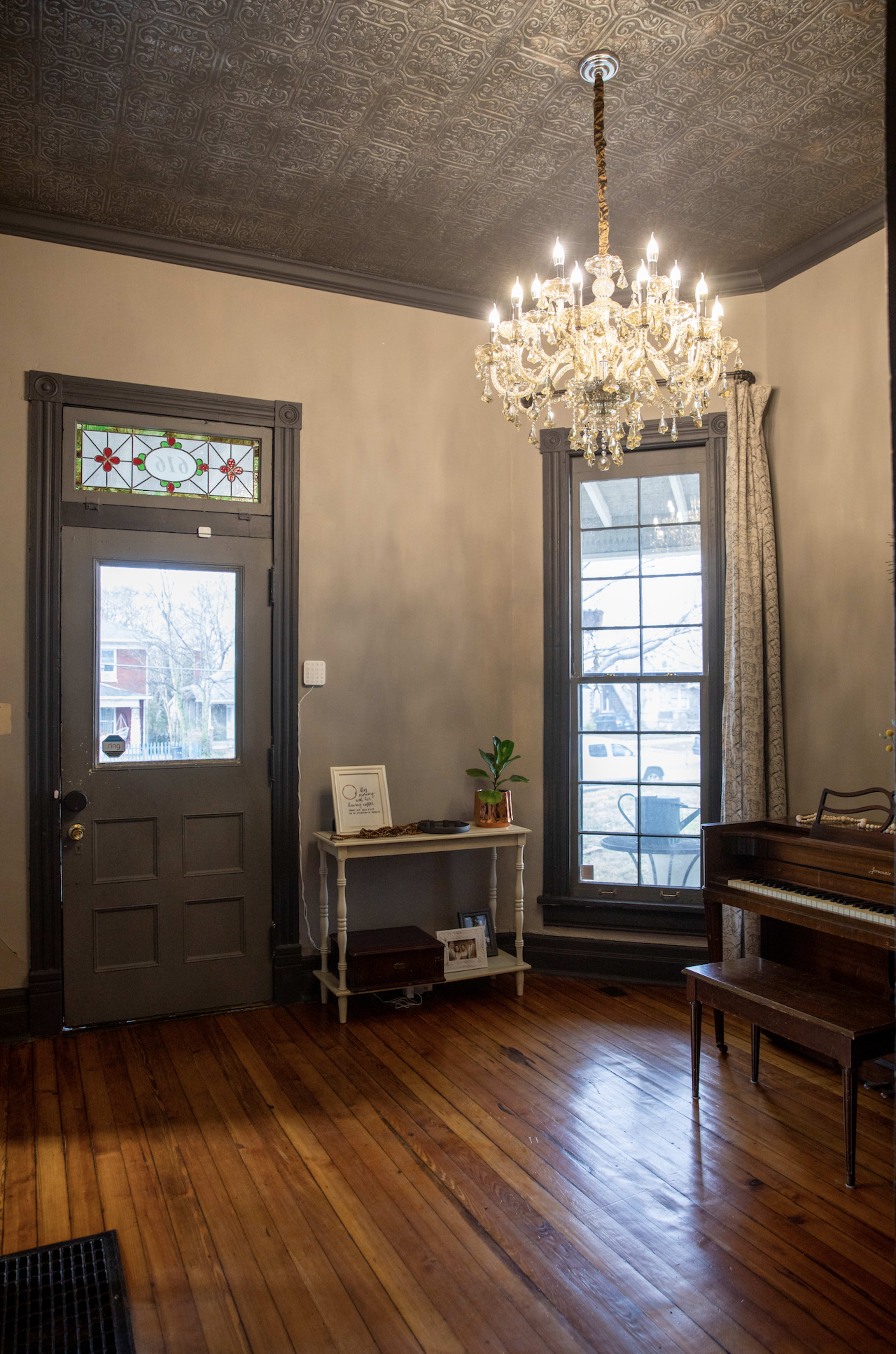 The image shows a room with a decorative ceiling, a chandelier, and a doorway that leads outside, featuring a piano and a small table against the wall.