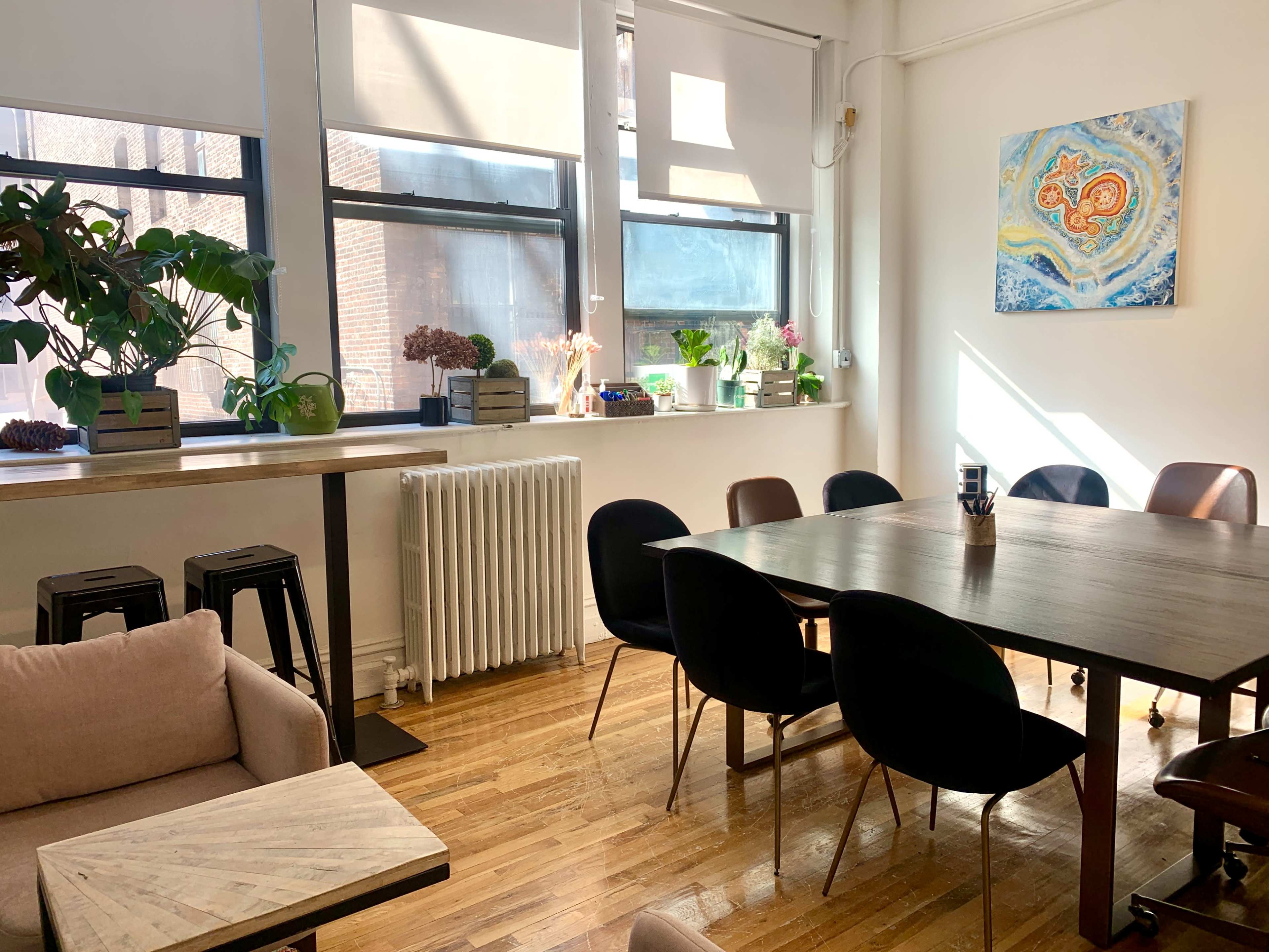 A bright, modern meeting room with a large wooden table, several chairs, plants, and artwork on the walls.