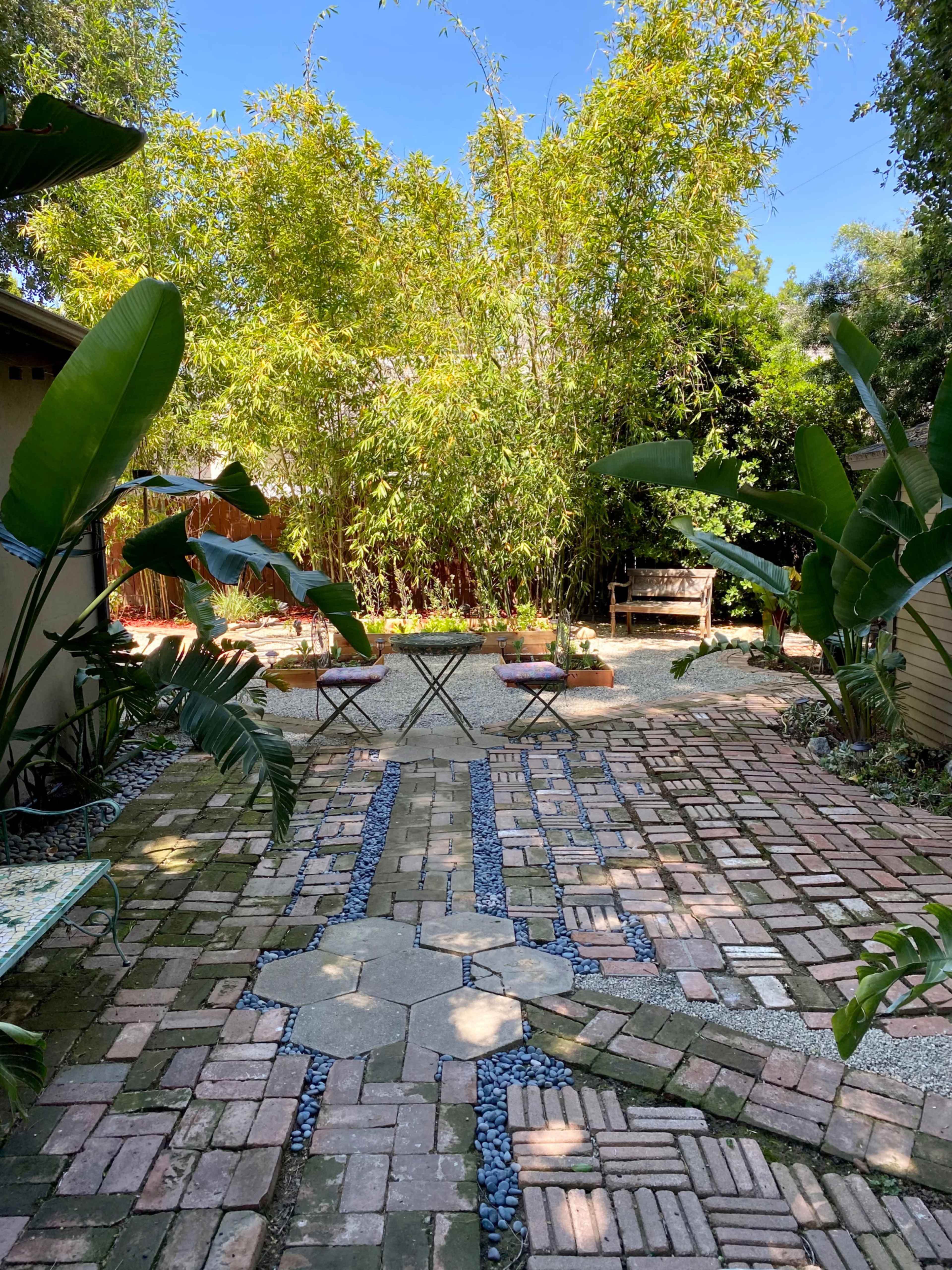 A landscaped garden with a brick pathway, bamboo plants, and seating areas surrounded by greenery.