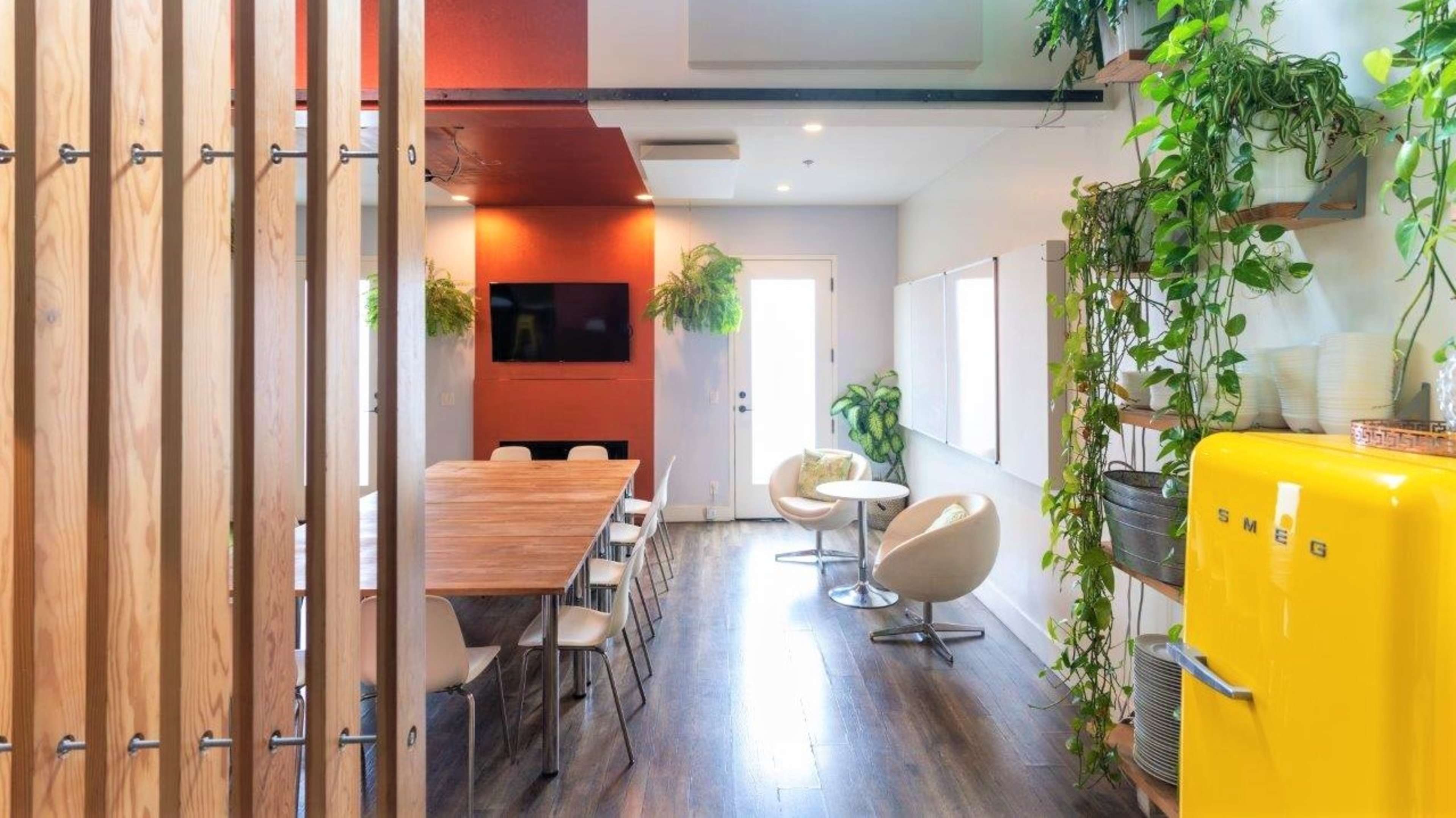 A bright, modern dining area features a large wooden table, round chairs, a yellow refrigerator, and various plants along the walls.