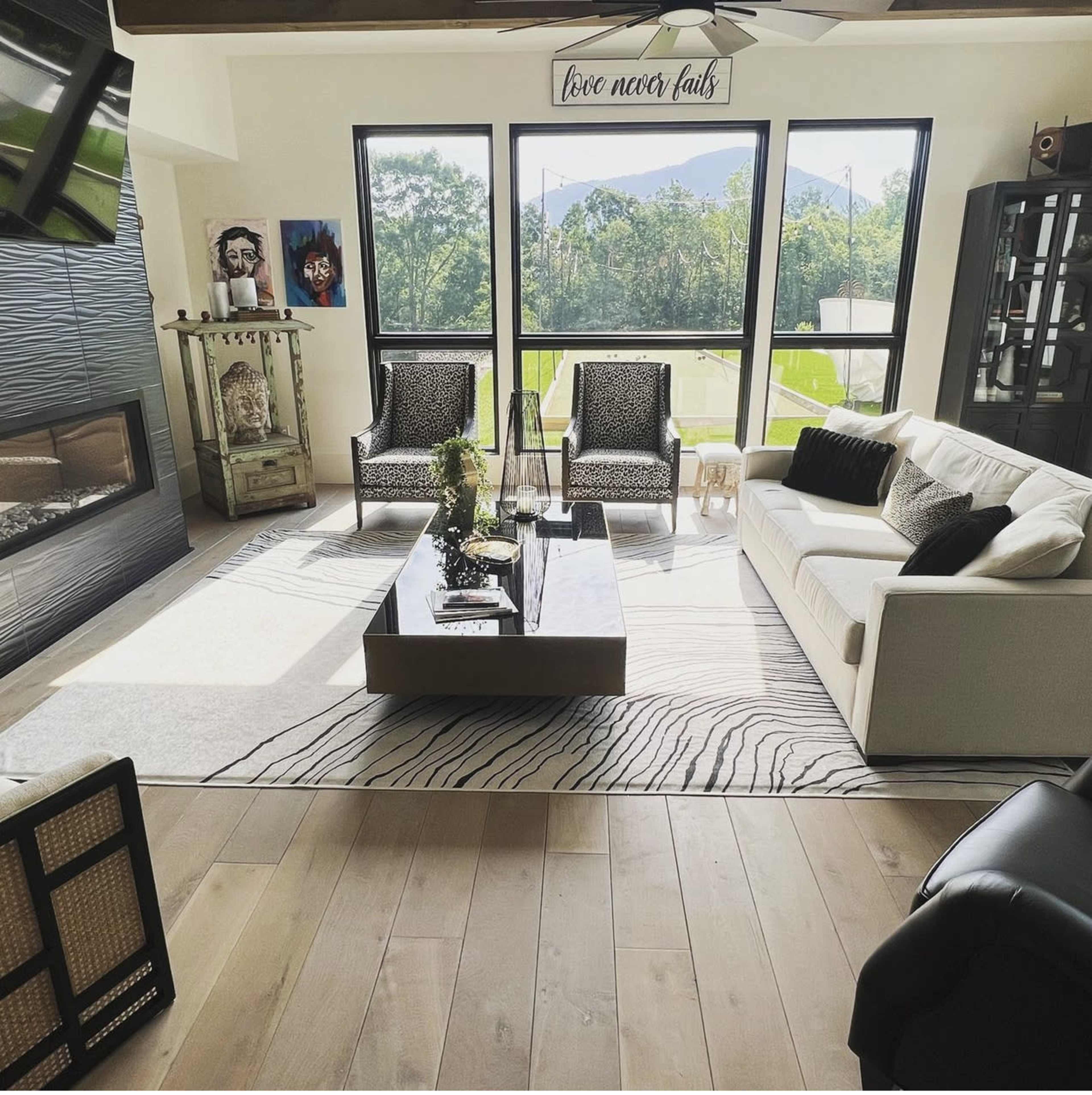 The living room features two patterned chairs, a white sofa, and a coffee table on a striped rug, with large windows that overlook a green landscape.