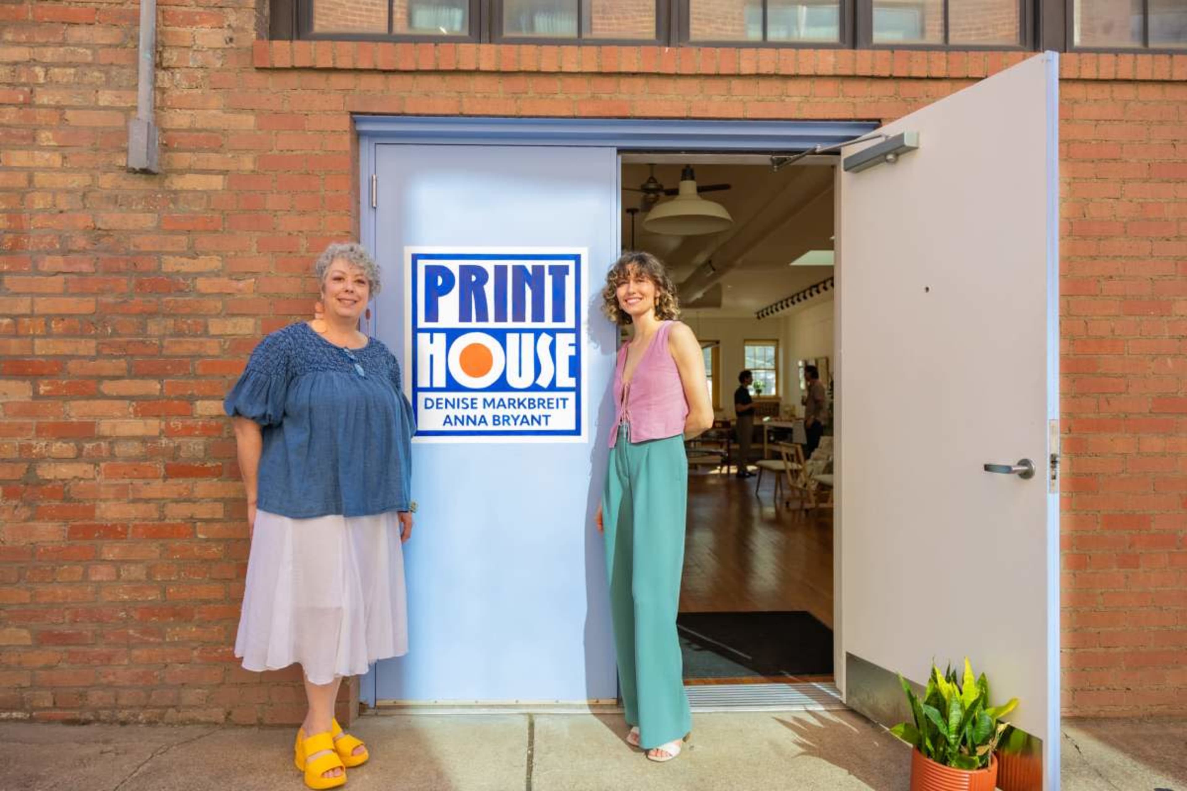 Two women stand at the entrance of a building labeled "Print House," one wearing a blue sweater and the other in a pink top and green pants.