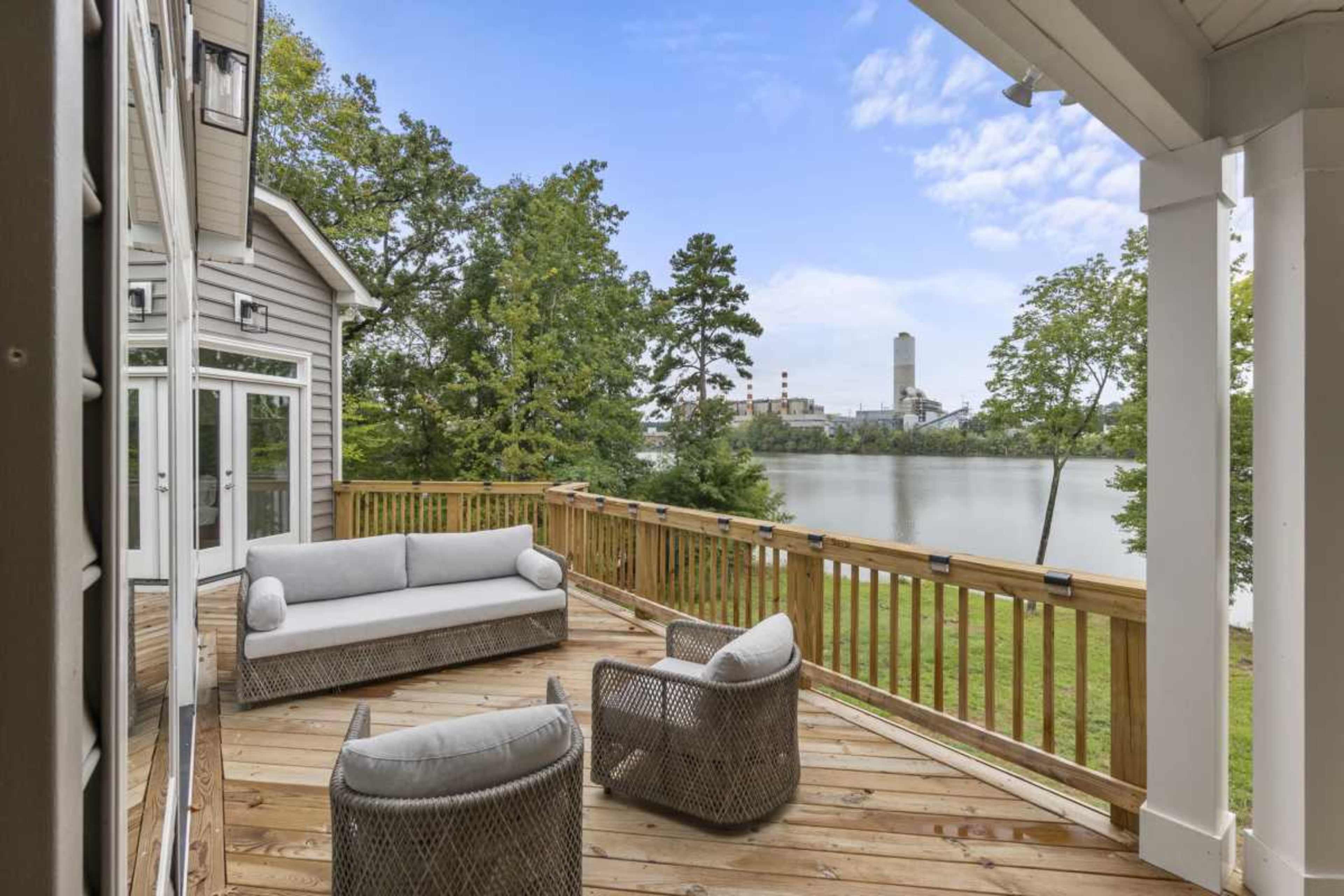 The image shows a wooden deck overlooking a river, featuring a gray sofa and two chairs, with trees and a building in the background.
