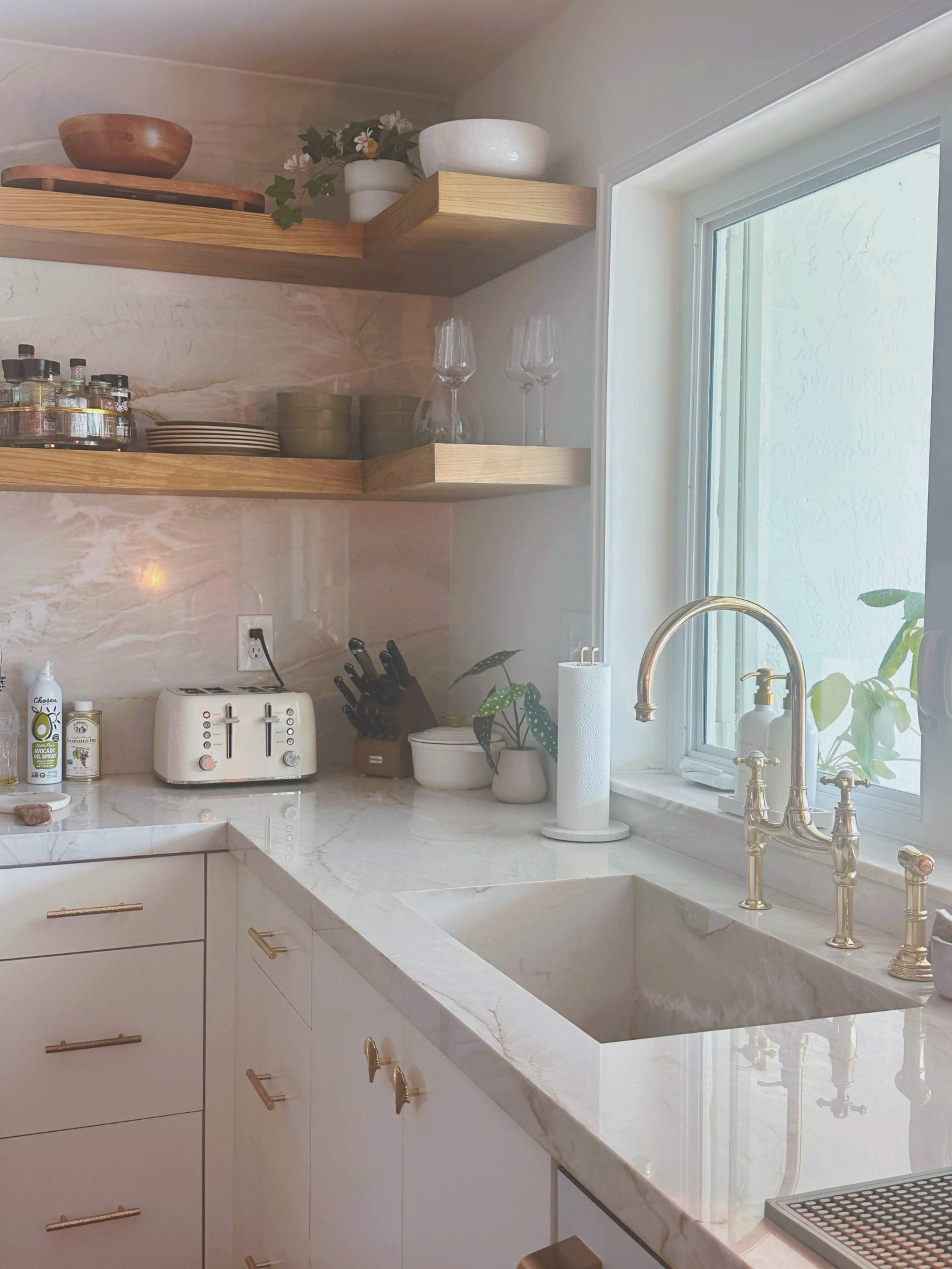 The kitchen features white cabinets, wooden shelves with various kitchen items, a marble countertop, and a window above the sink.