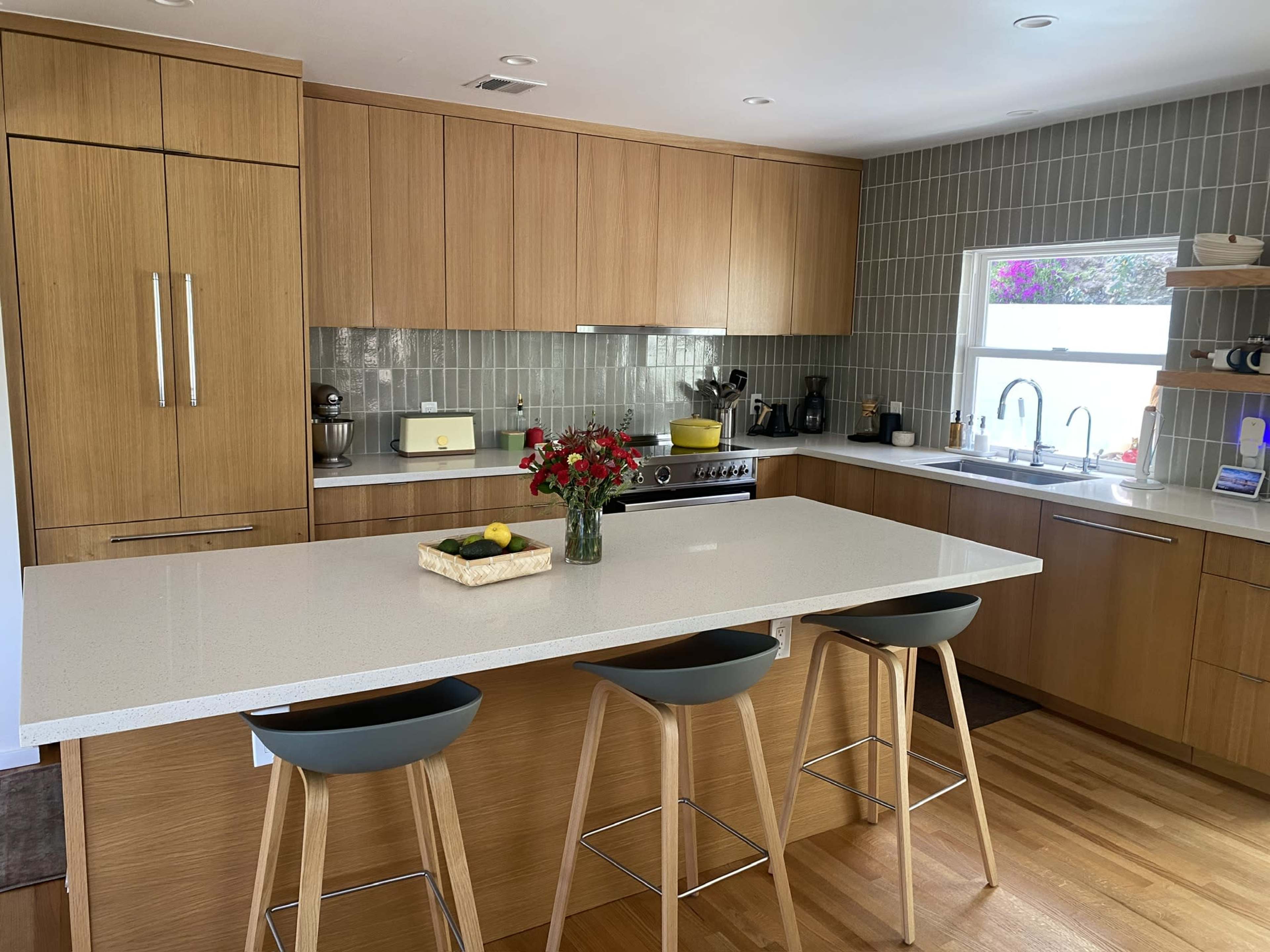 The image shows a modern kitchen with wooden cabinets, a white countertop with a vase of flowers, and three stools at the island.