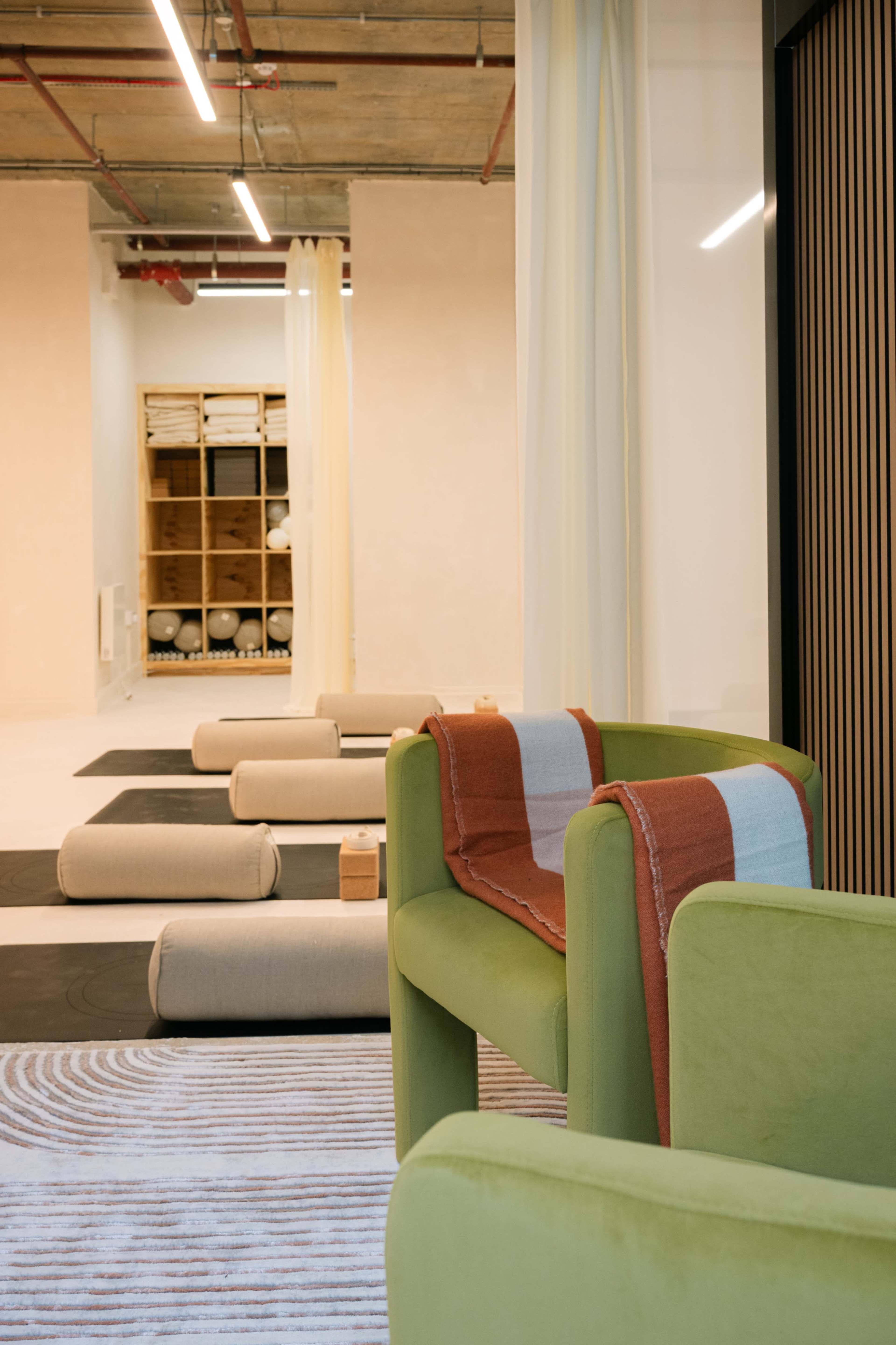 The image shows a modern interior space featuring green armchairs, rolled mats, and a storage area in the background.