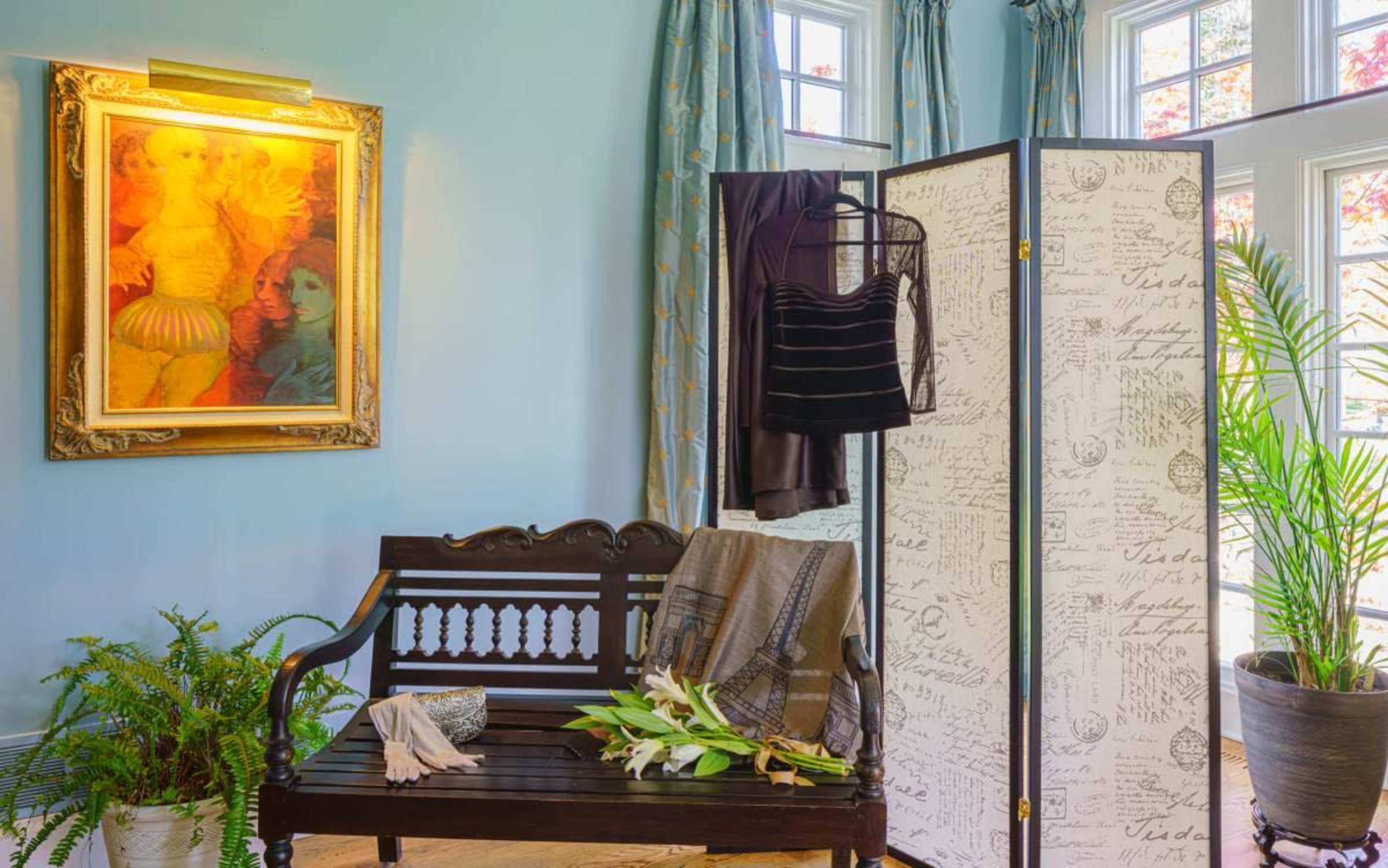 A wooden bench is placed in a well-lit room featuring a folding screen, a decorative painting on the wall, and various clothing items displayed nearby.