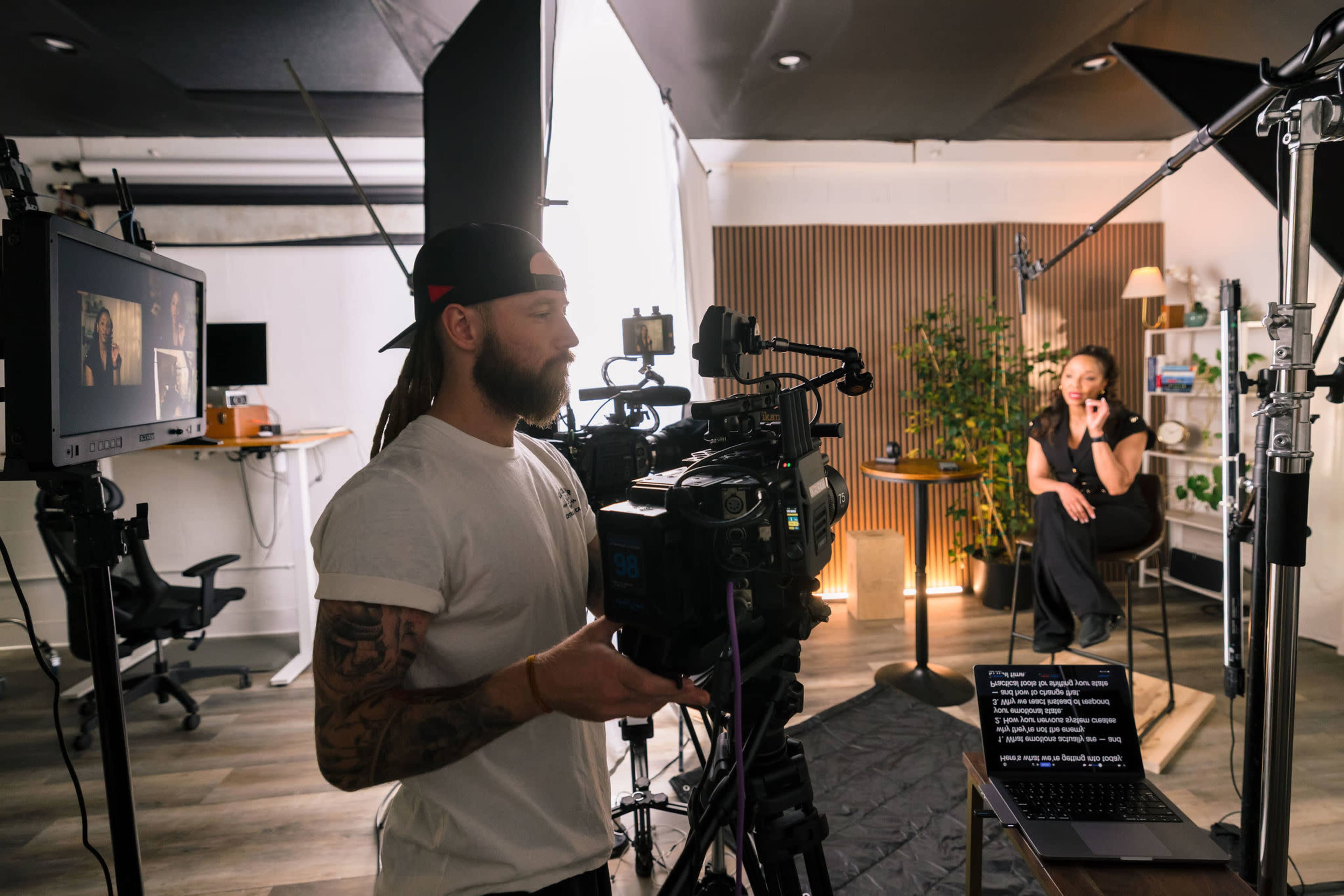 A cameraman operates a video camera while a woman sits in front of a backdrop, engaging in an interview setup with lighting and equipment arranged around them.