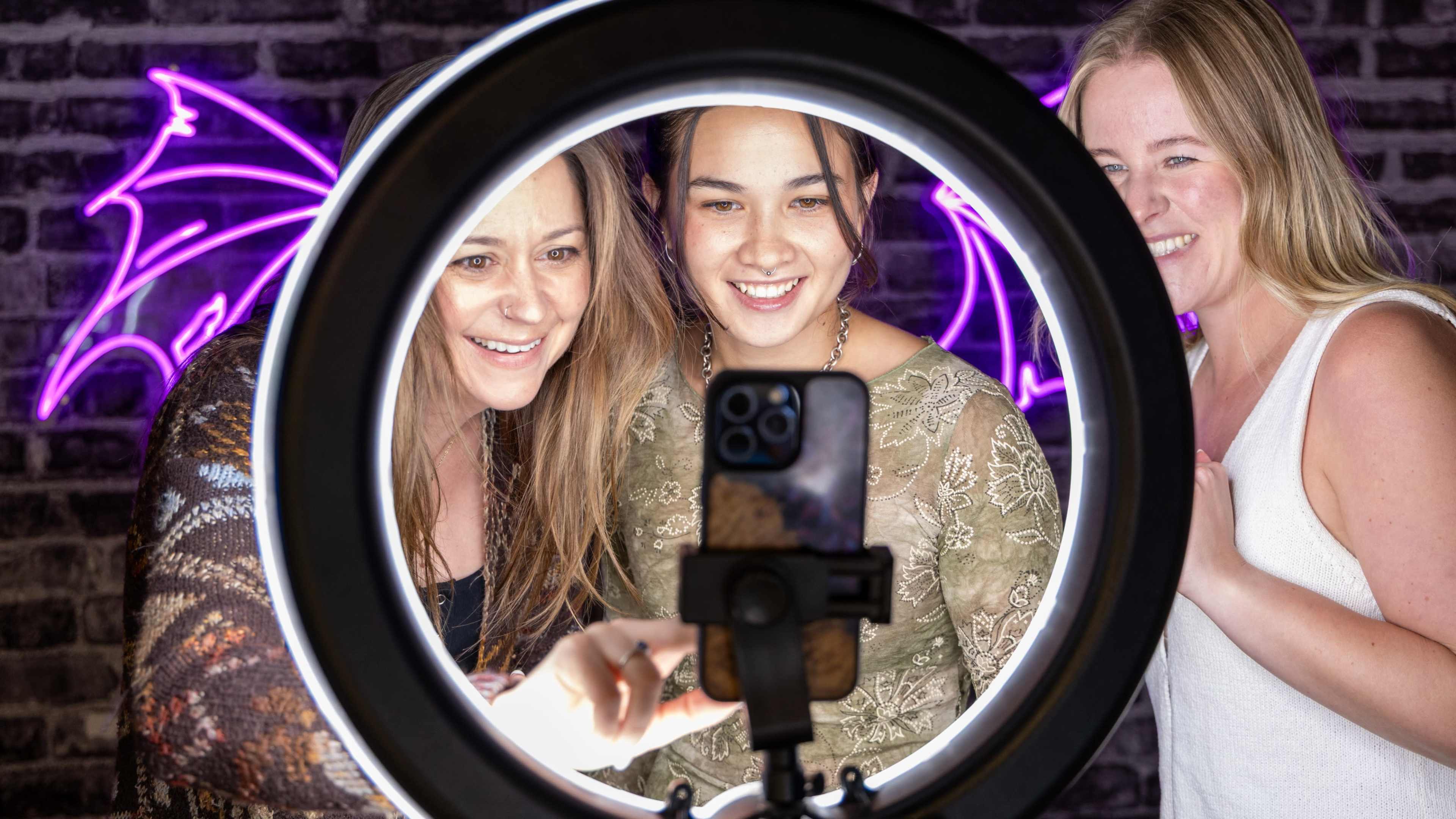 Three women smile while taking a selfie together using a smartphone mounted in a ring light.