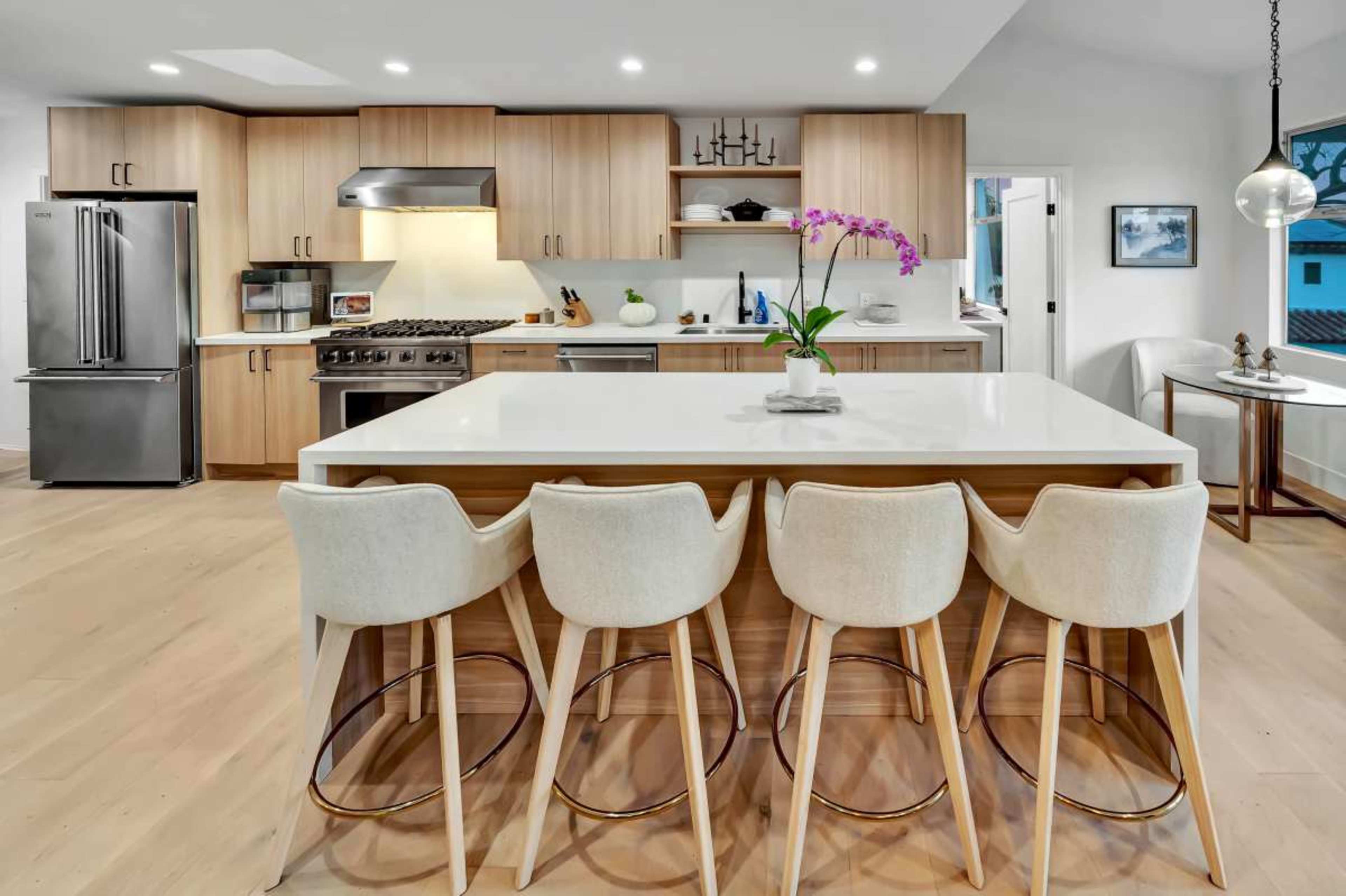 A modern kitchen features light wood cabinetry, stainless steel appliances, and a large central island with four barstools.