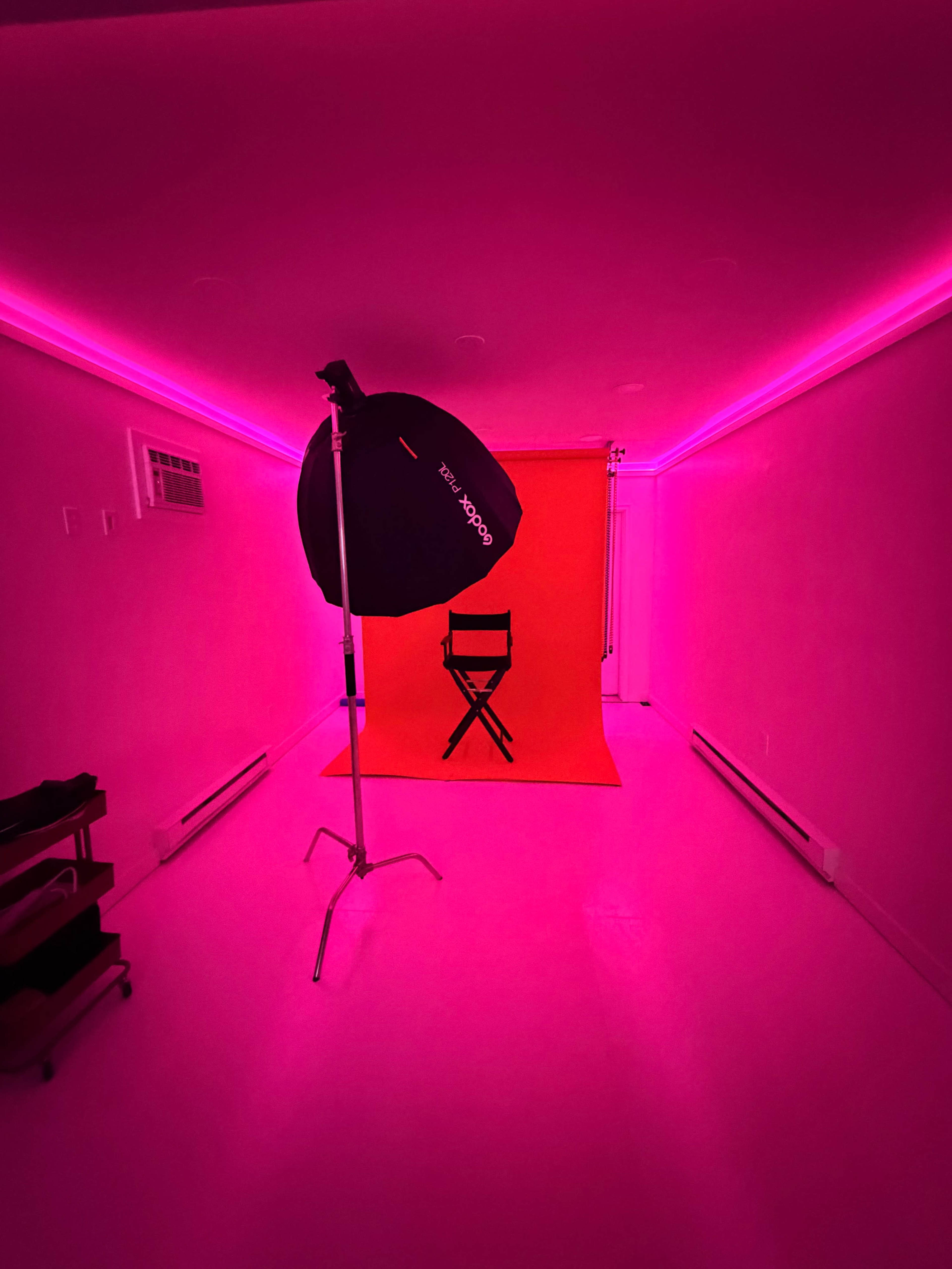 A photography studio is illuminated with pink lighting, featuring a director's chair against a bright pink backdrop.