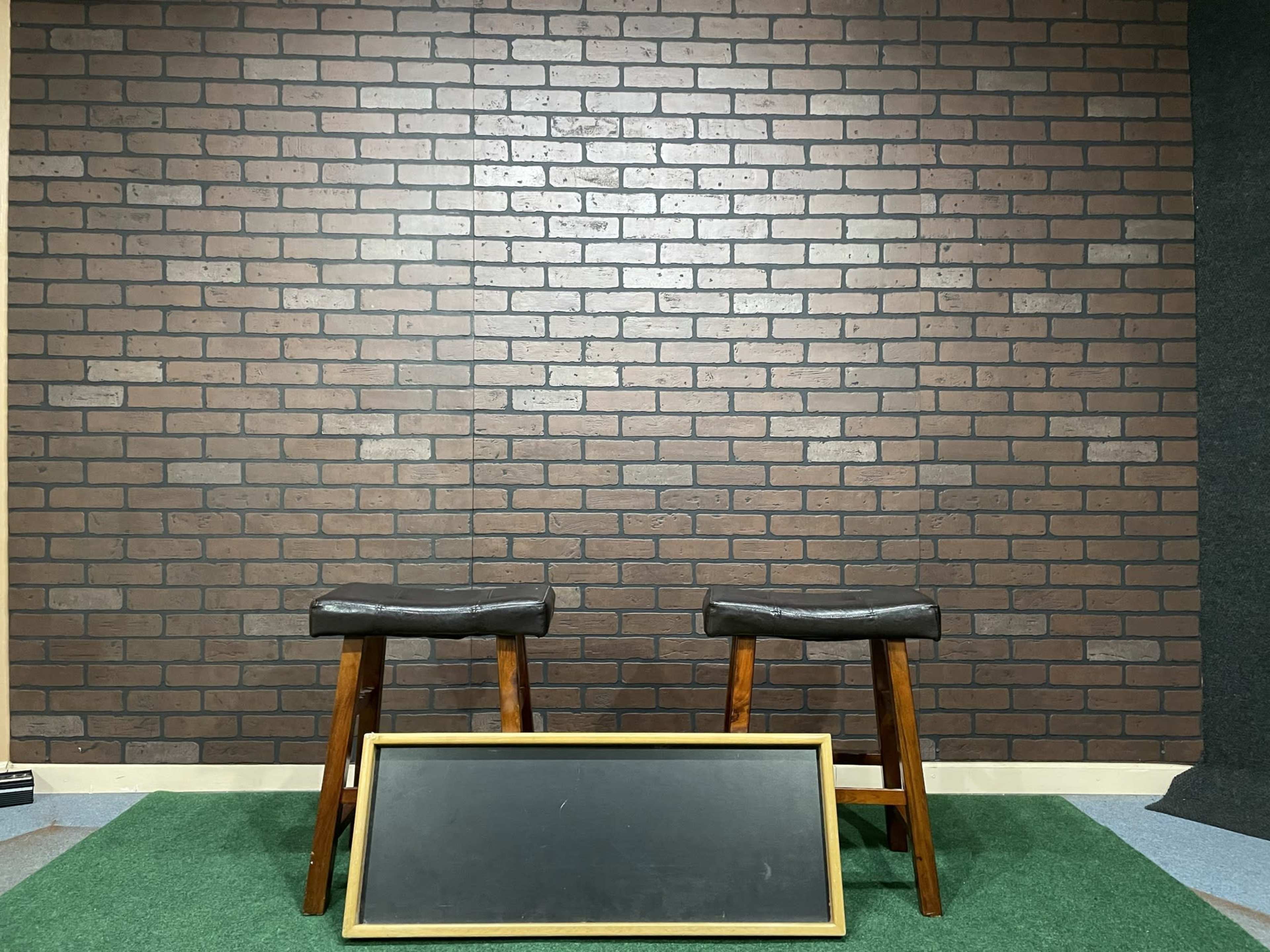 Two wooden stools with black seats positioned in front of a brick-patterned wall, with a blank blackboard leaning against the floor.