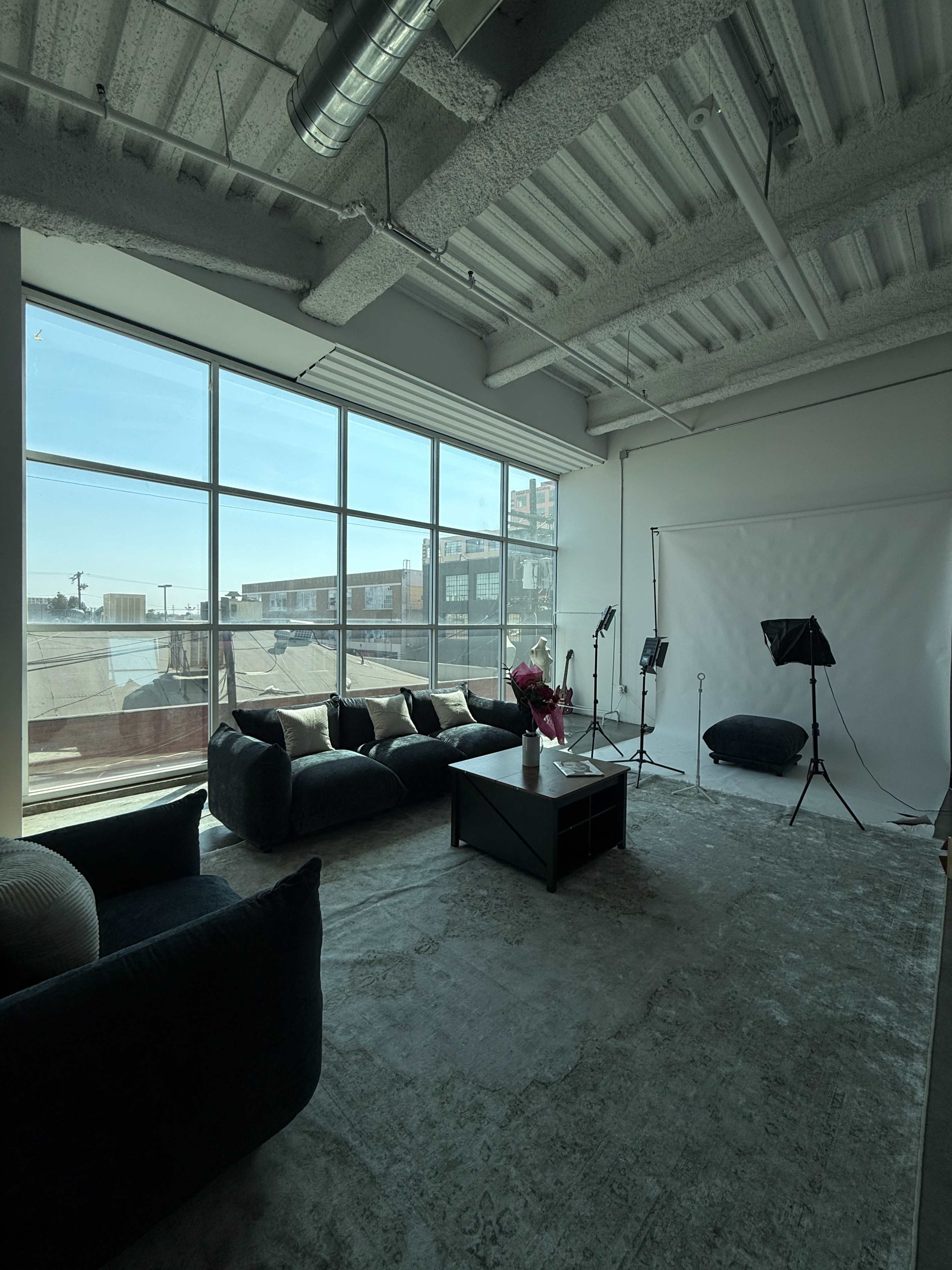 The image shows a modern indoor space with large windows, a dark sofa set, a coffee table, and studio lighting equipment.