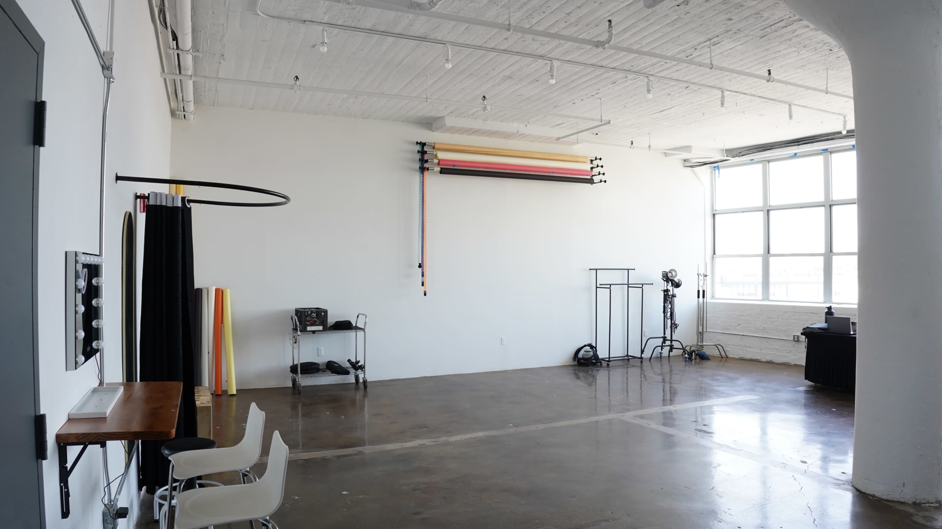 The image shows a spacious, empty studio with white walls, polished concrete flooring, and equipment such as backdrop stands and lights along one side.