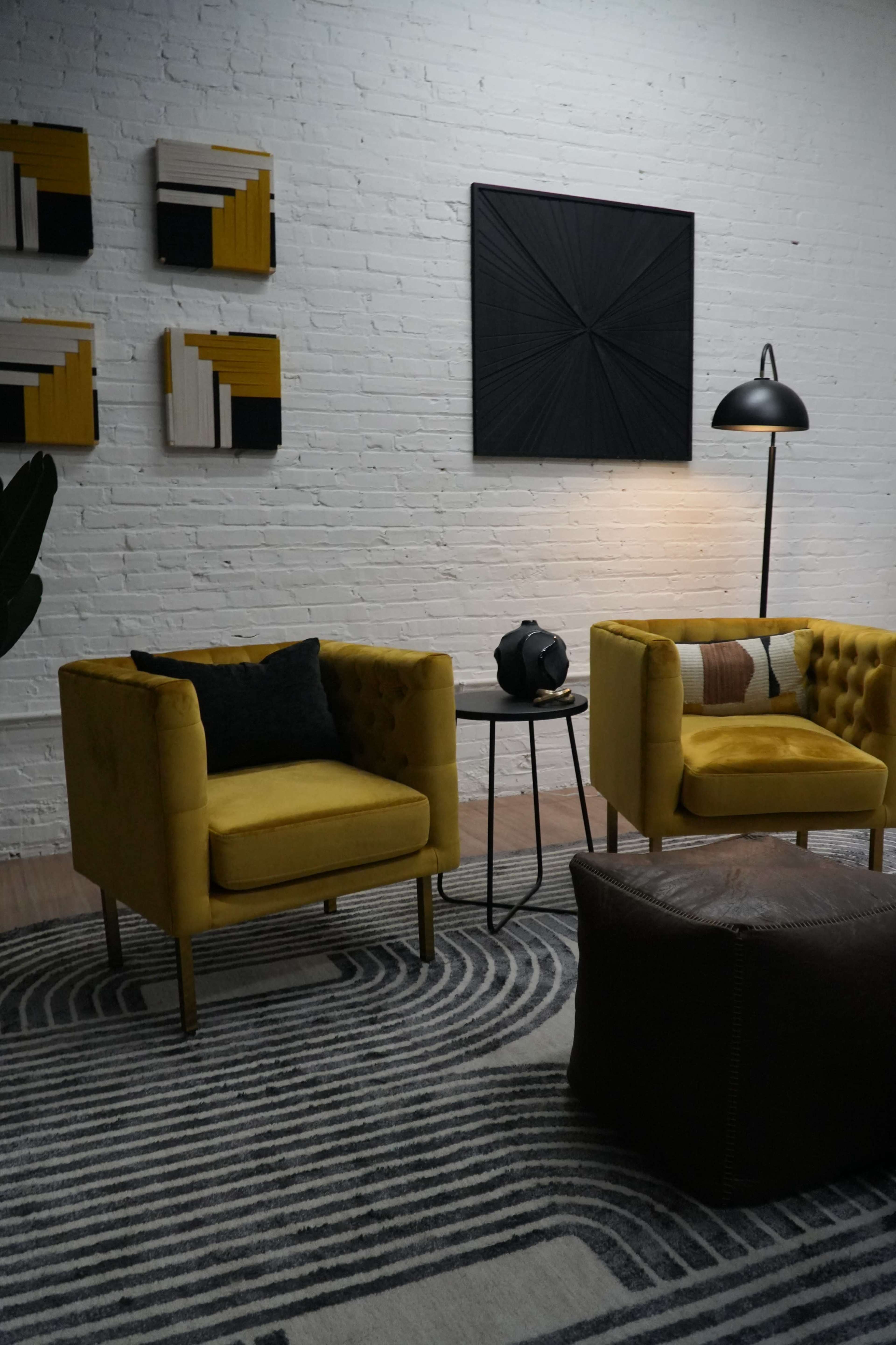 The image shows a modern living room with two yellow upholstered armchairs, a round side table, a patterned area rug, and abstract wall art above them.