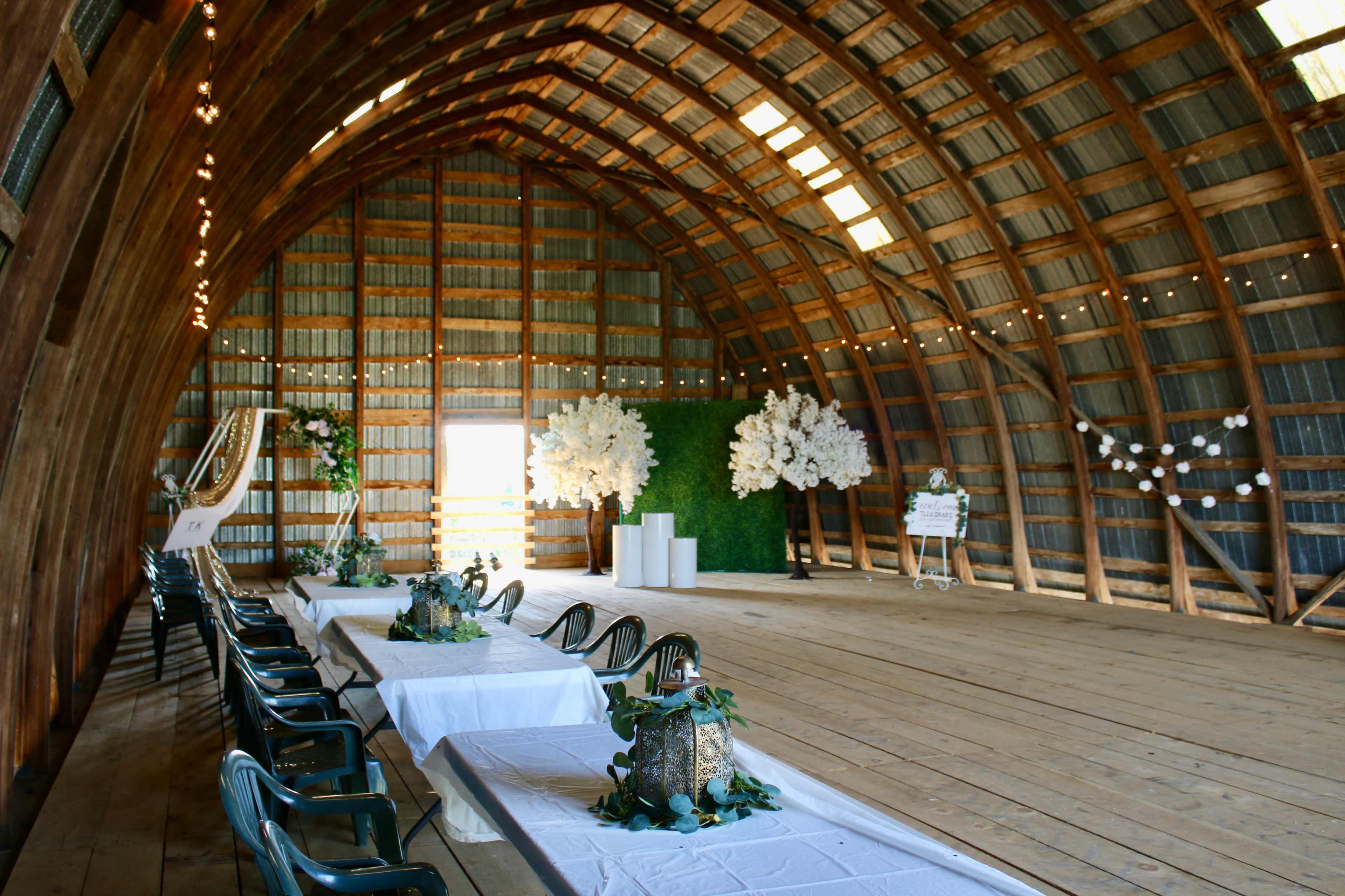 A spacious wooden barn interior set up for an event, featuring a long table with decorations, chairs, and a decorative backdrop of white flowers and greenery.
