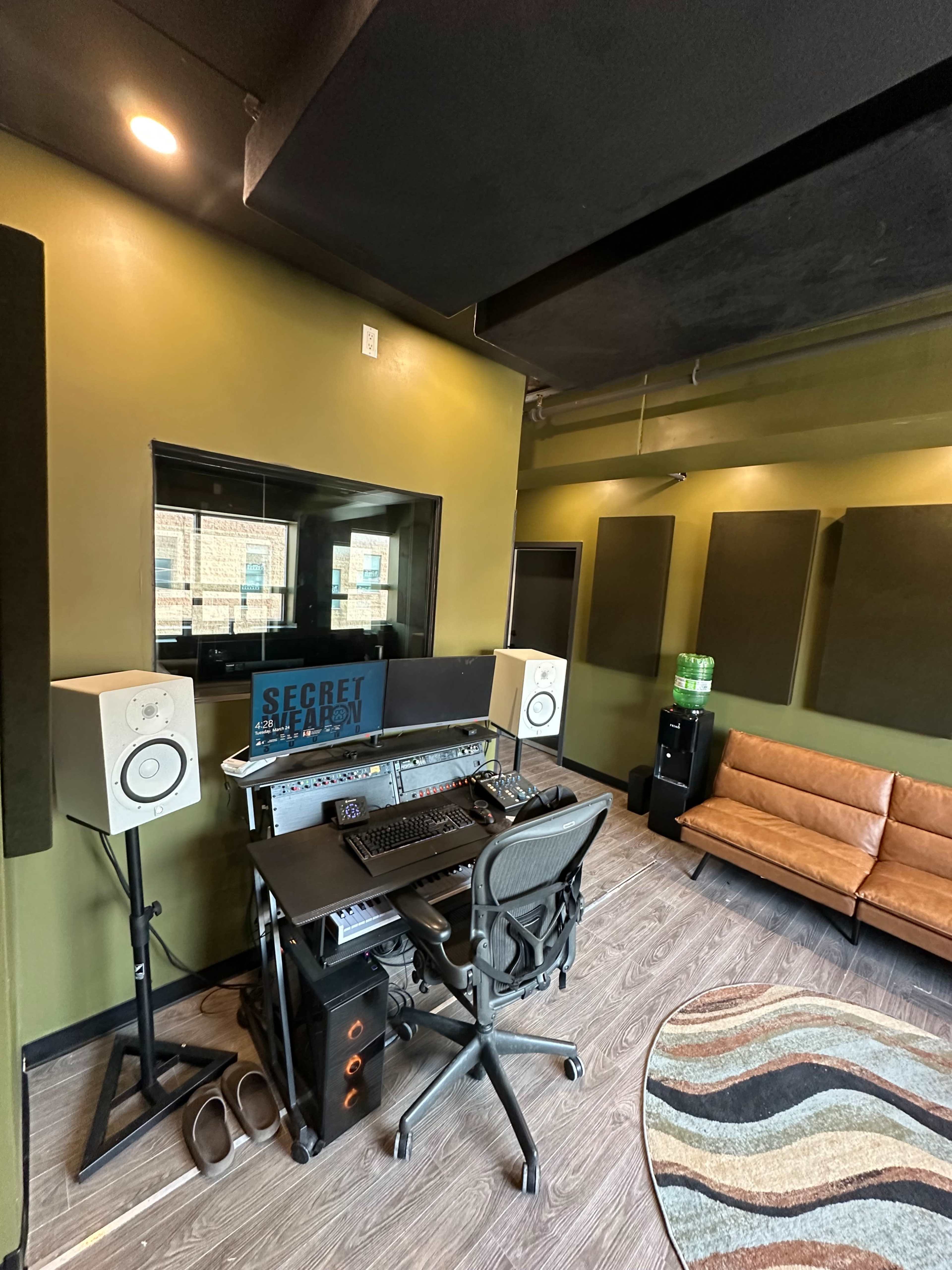 The image shows a recording studio with a control desk, speakers, a chair, and a brown sofa against green walls.