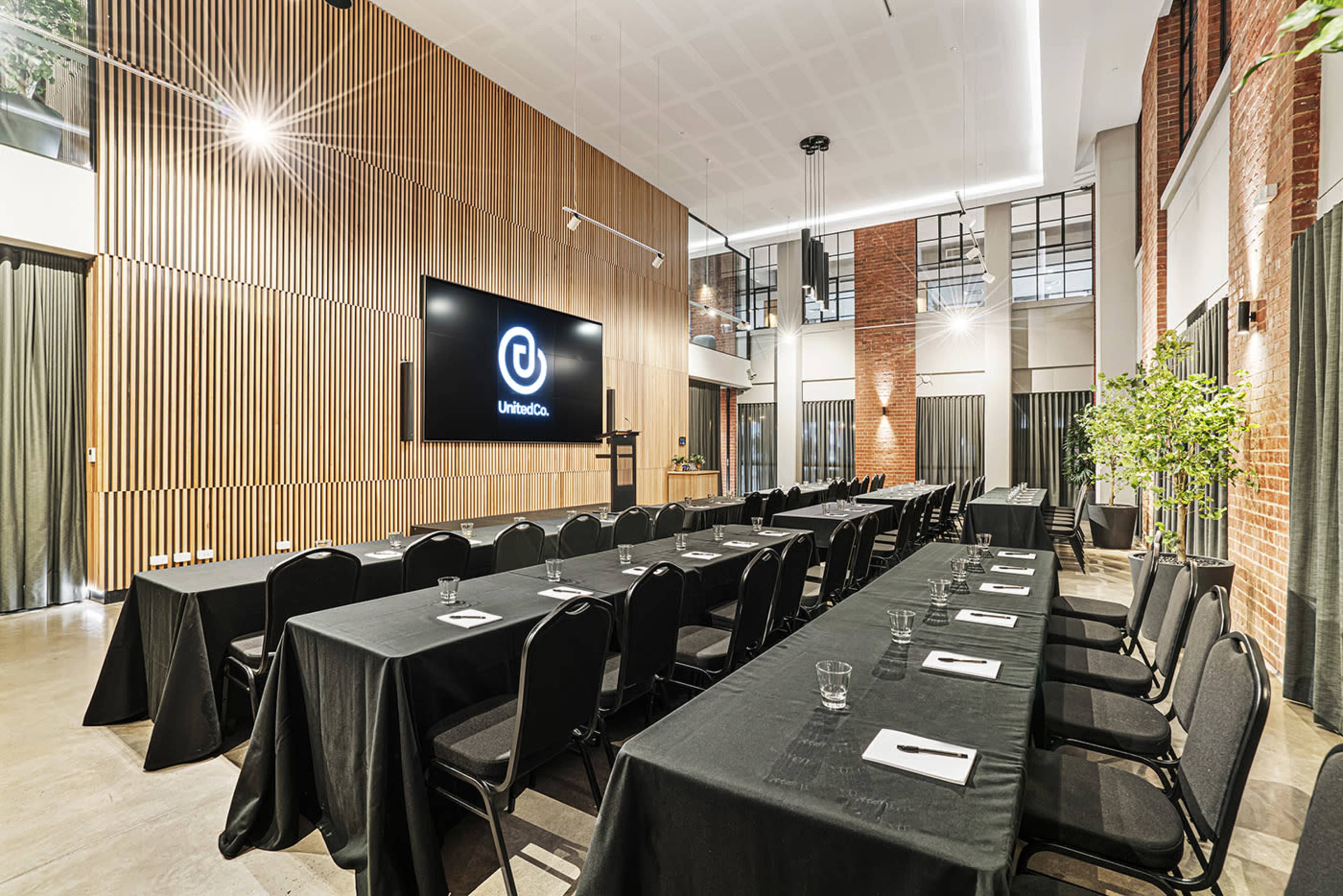 The image shows a modern conference room with rows of black tables and chairs arranged facing a large screen on a wooden accent wall.