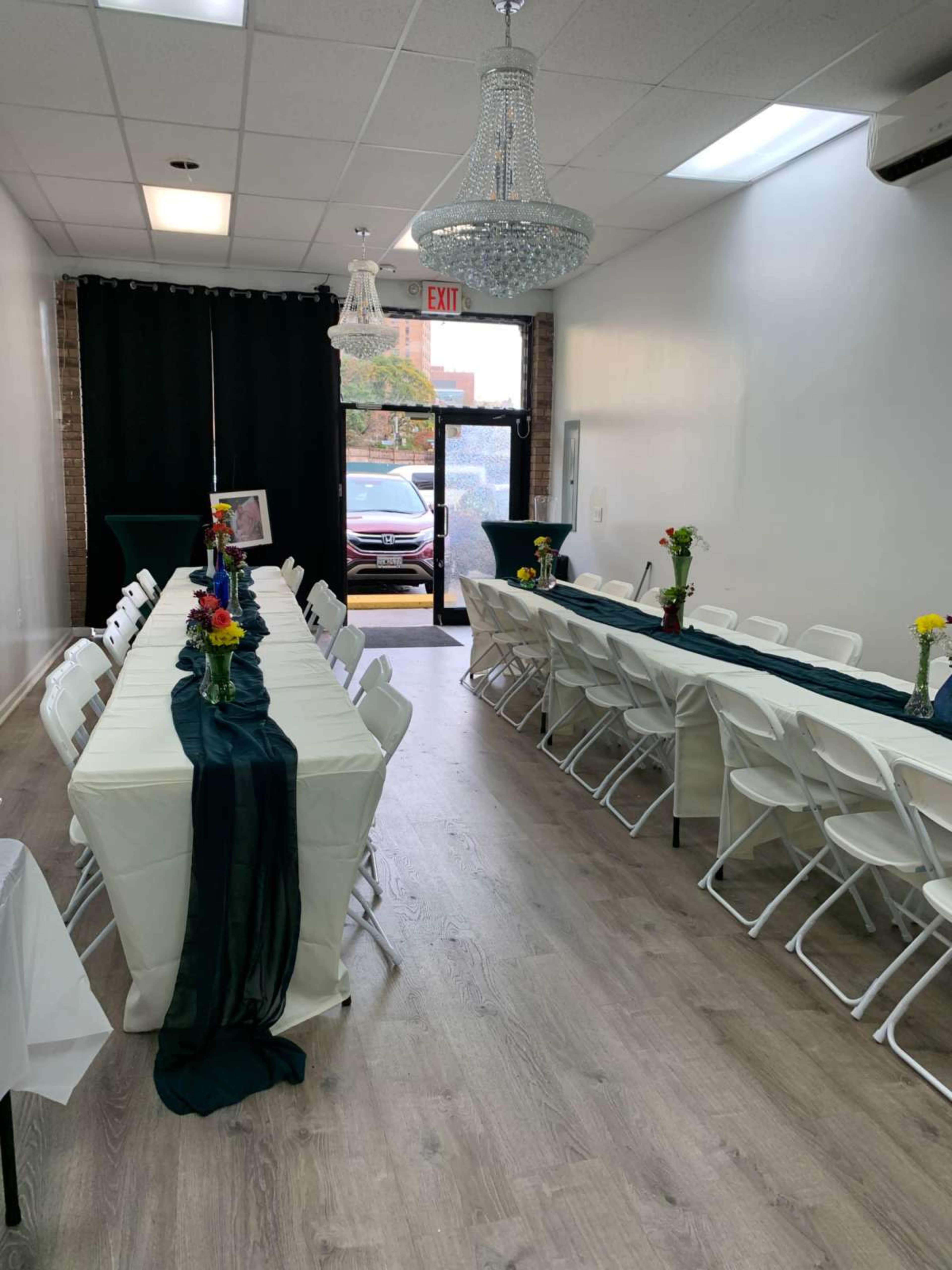 The image shows a bright event space featuring two long tables covered with white tablecloths, adorned with centerpieces of flowers, and illuminated by two chandeliers.