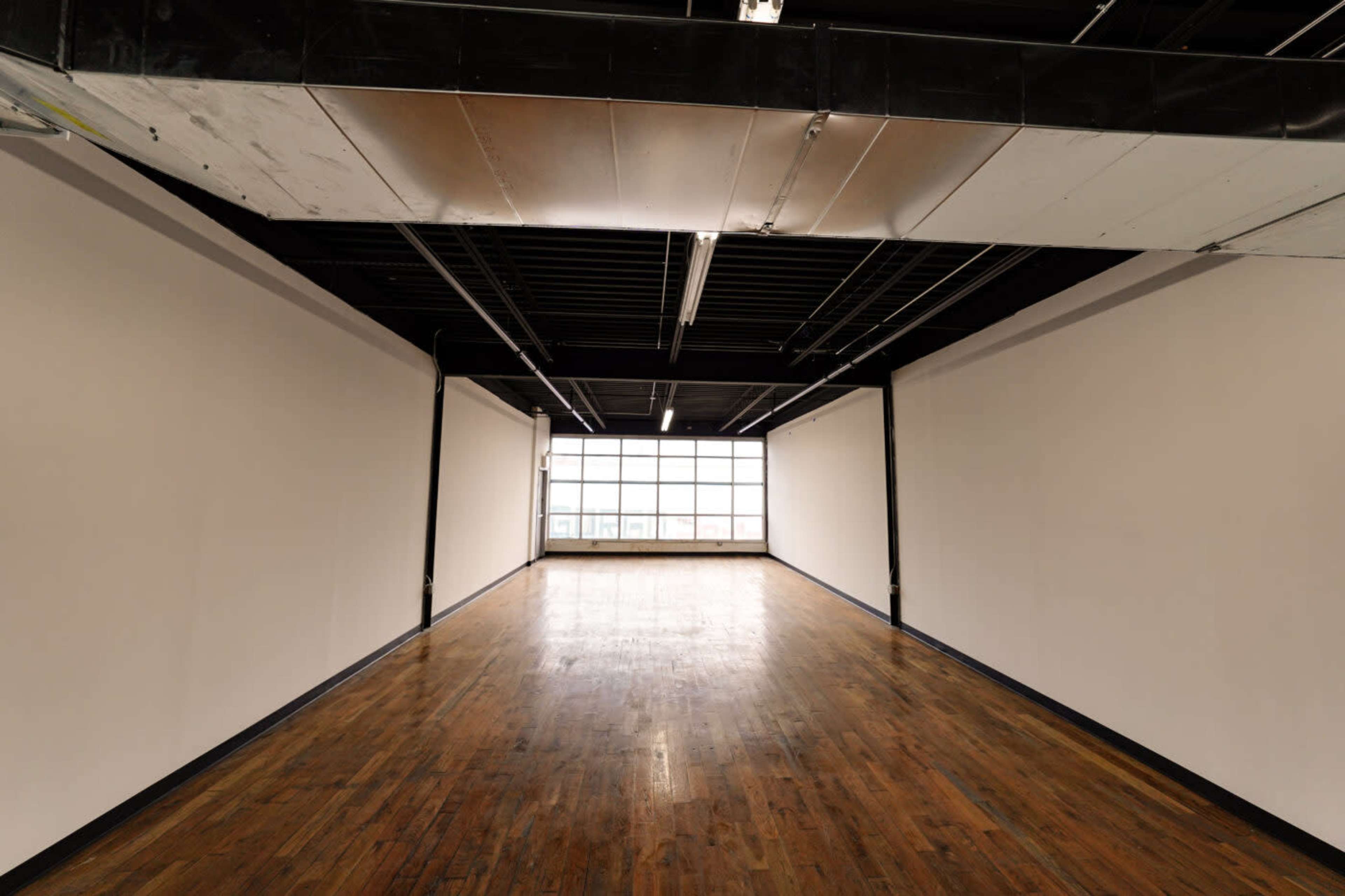 The image shows an empty room with wooden flooring and large windows at one end, surrounded by plain walls.